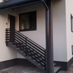 Zewnętrzne schody z ciemną, metalową balustradą prowadzące do drzwi wejściowych w jasnym budynku. Widoczny fragment elewacji i okna z żaluzjami.