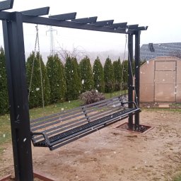 STEEL TEAM Strzałka Piotr - Czarna, metalowa huśtawka ogrodowa z łańcuchami, w tle rząd zielonych tuj i szklarnia, podczas opadów śniegu.