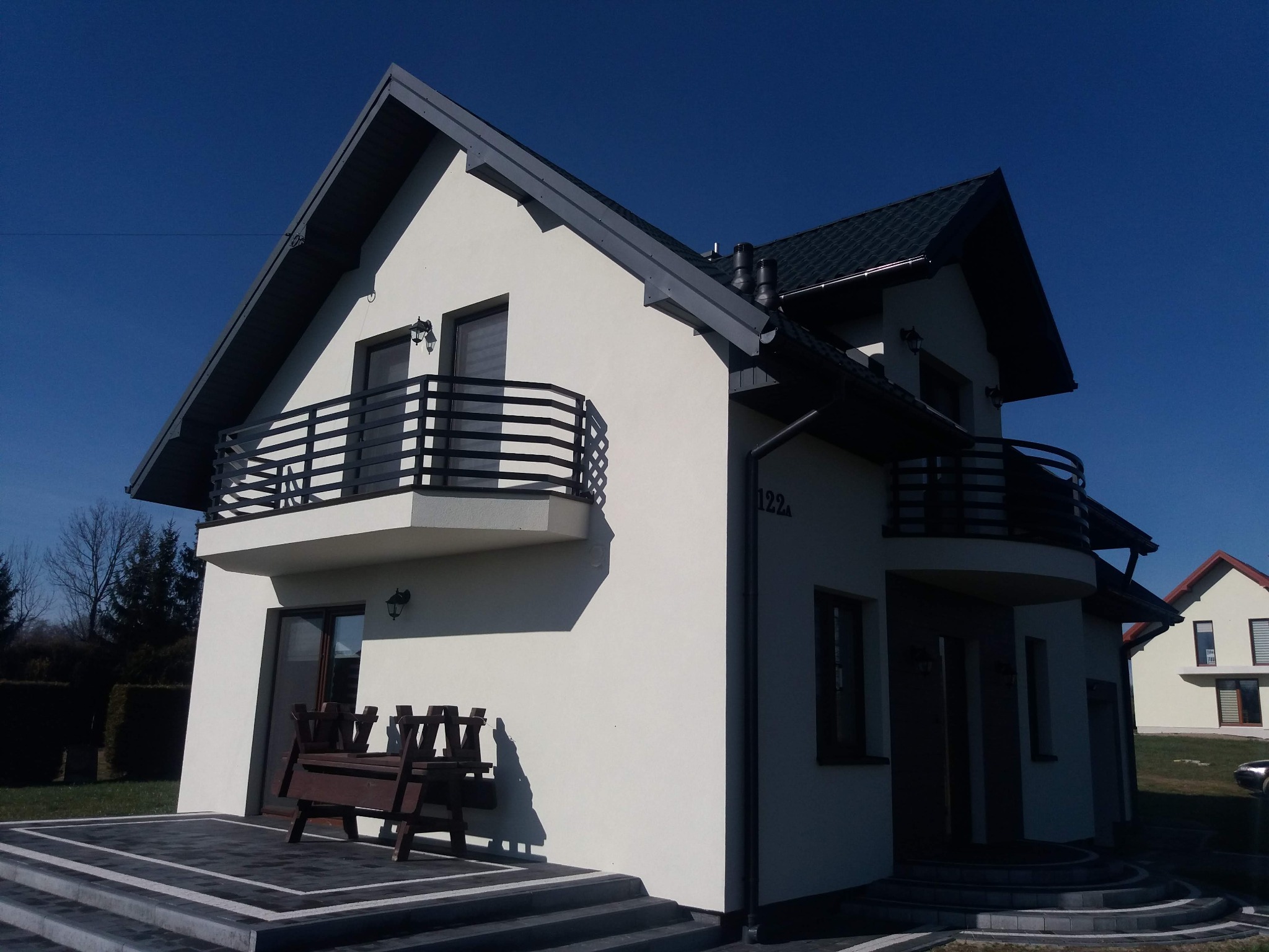 Nowoczesny dom z balkonem z czarną, poziomą balustradą i numerem 122a, w słoneczny dzień na tle błękitnego nieba. Widoczny fragment tarasu z ławką.
