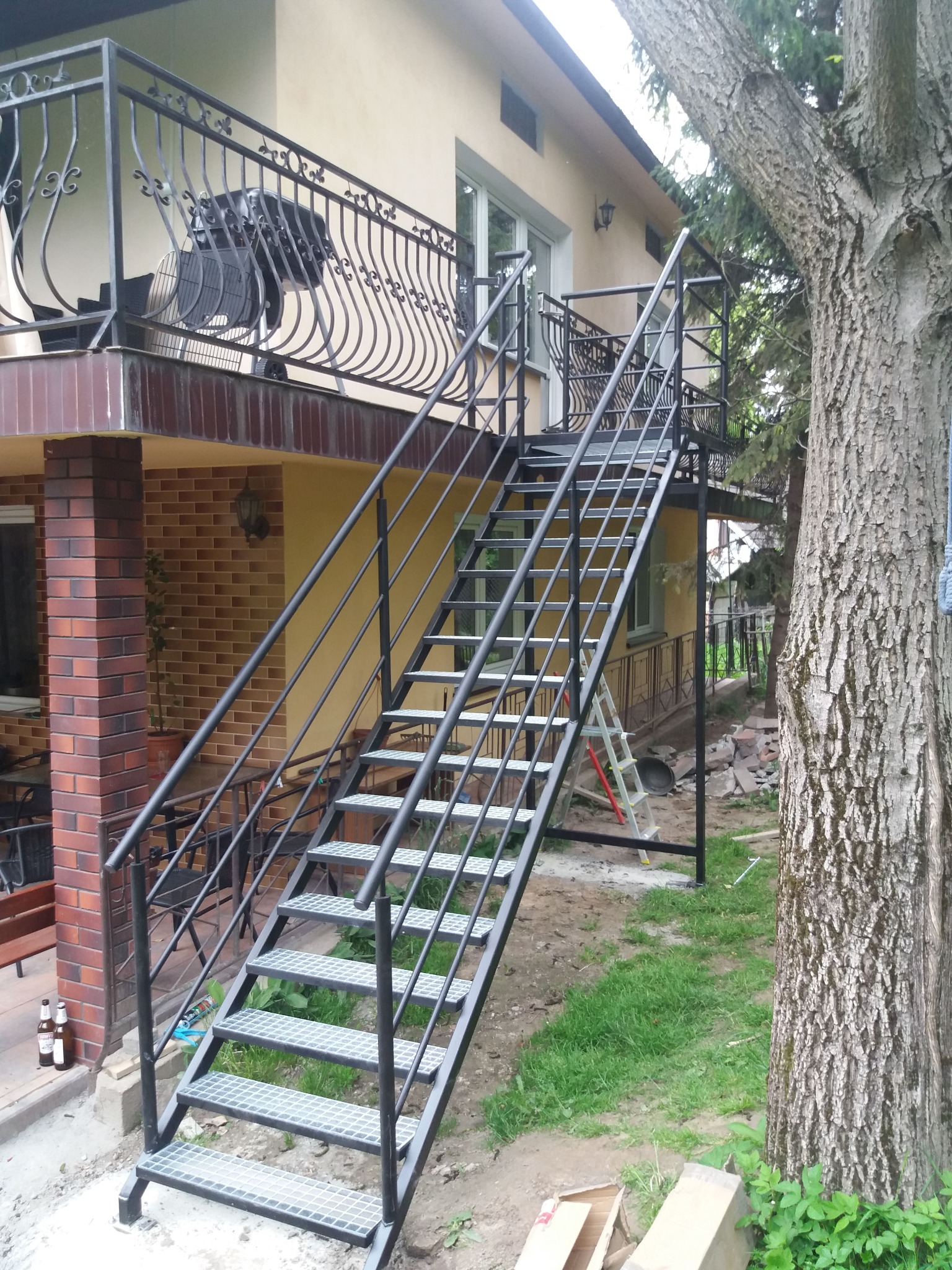 Zewnętrzne schody metalowe z balustradą prowadzące na balkon domu z beżową elewacją. Widoczne elementy wykończeniowe i duży pień drzewa obok.