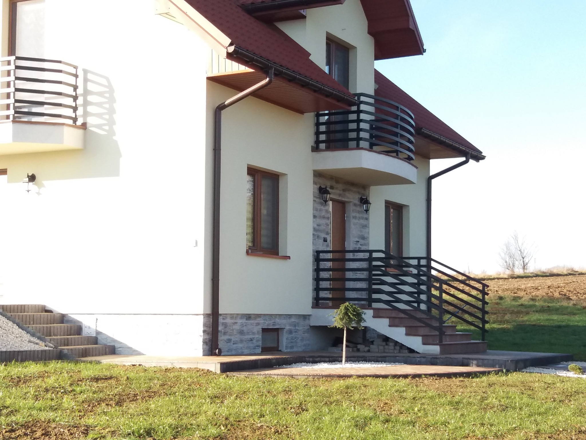 Nowoczesna balustrada balkonowa i schodowa z czarnych, metalowych profili przy domu jednorodzinnym w Ropczycach. Jasna elewacja, brązowe okna i dach.