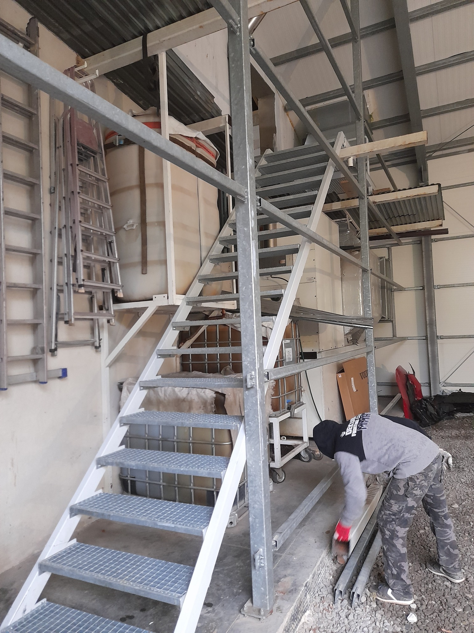 Metalowe schody przemysłowe z kratowymi stopniami, w trakcie montażu, z pracownikiem w odzieży roboczej. Widoczne elementy konstrukcyjne i drabiny w tle.
