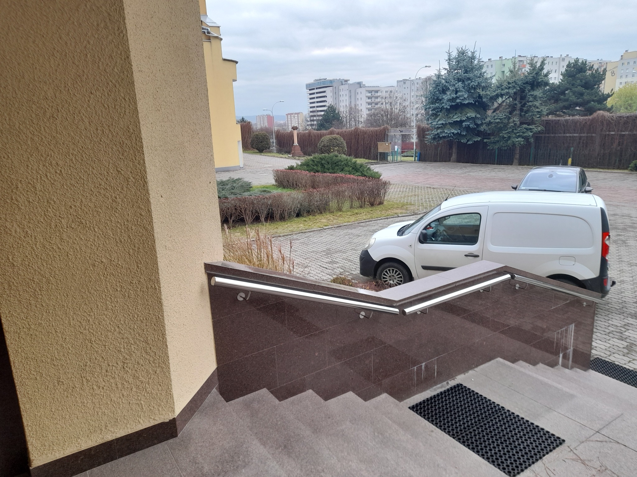 Zewnętrzne schody z brązowego granitu z poręczą ze stali nierdzewnej, widok z góry. W tle budynki i zieleń miejska w pochmurny dzień.