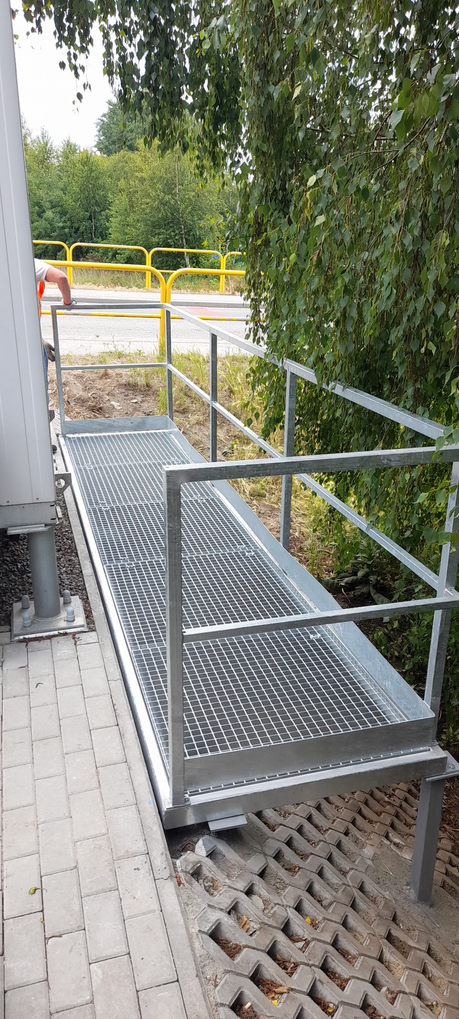 Metalowa kładka z kratą i barierką, łącząca budynek z terenem. Widoczne elementy konstrukcyjne, kostka brukowa i roślinność w tle. Szara kolorystyka.