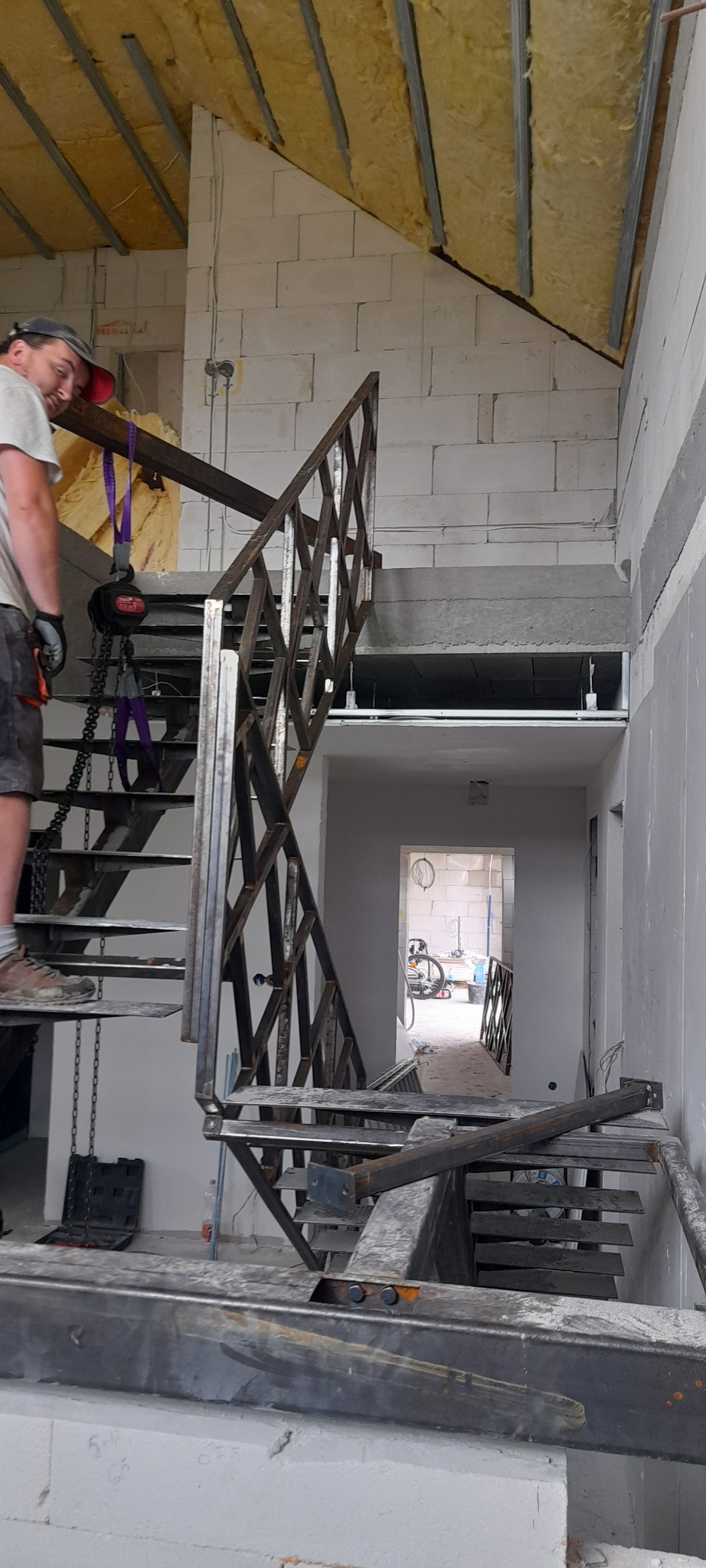 Montaż metalowej konstrukcji schodów w surowym wnętrzu. Widoczne elementy mocujące, pracownik w tle i dalszy plan z otwartym przejściem.