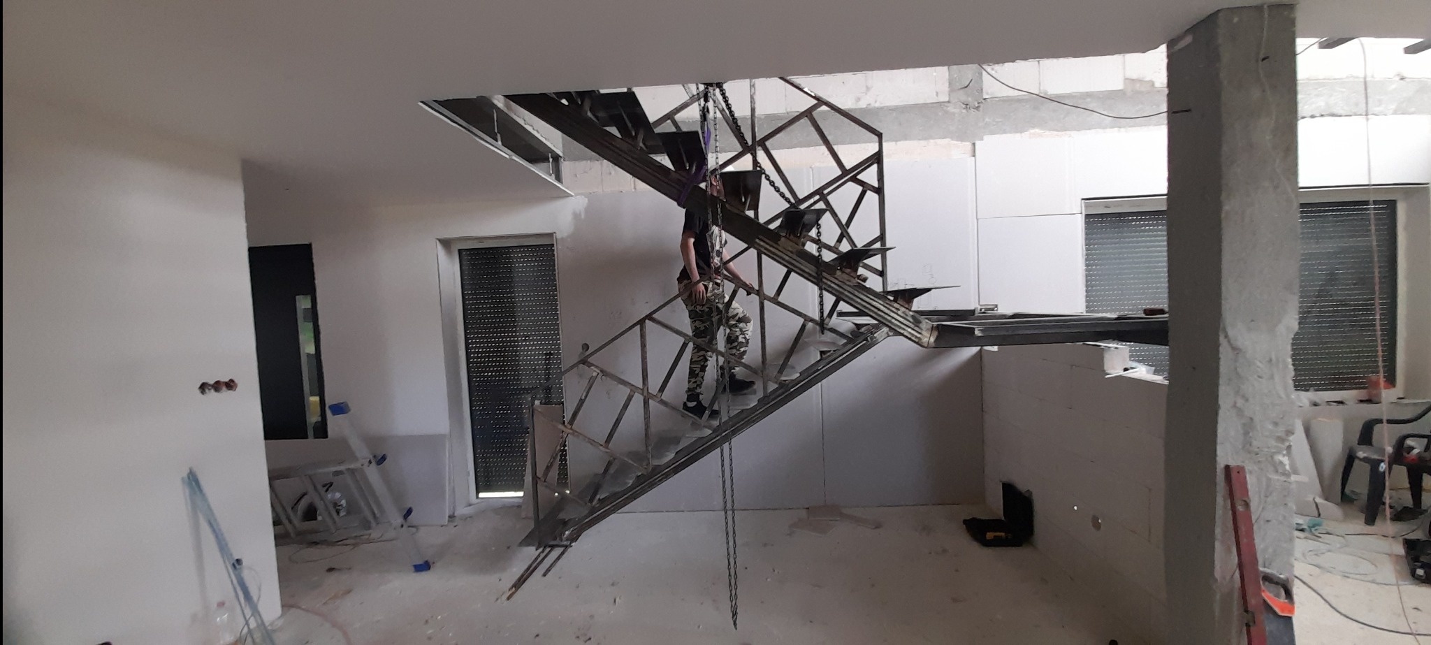 Montaż metalowych schodów w surowym wnętrzu. Konstrukcja podwieszona na łańcuchach, widoczny pracownik w trakcie instalacji. W tle okna z żaluzjami.