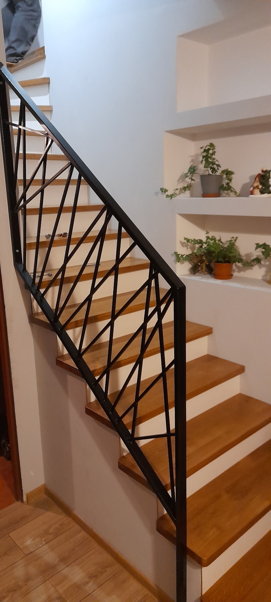 Nowoczesne schody z drewnianymi stopniami i czarną, geometryczną balustradą. W tle białe ściany z półkami i roślinami doniczkowymi. Ujęcie z dołu.