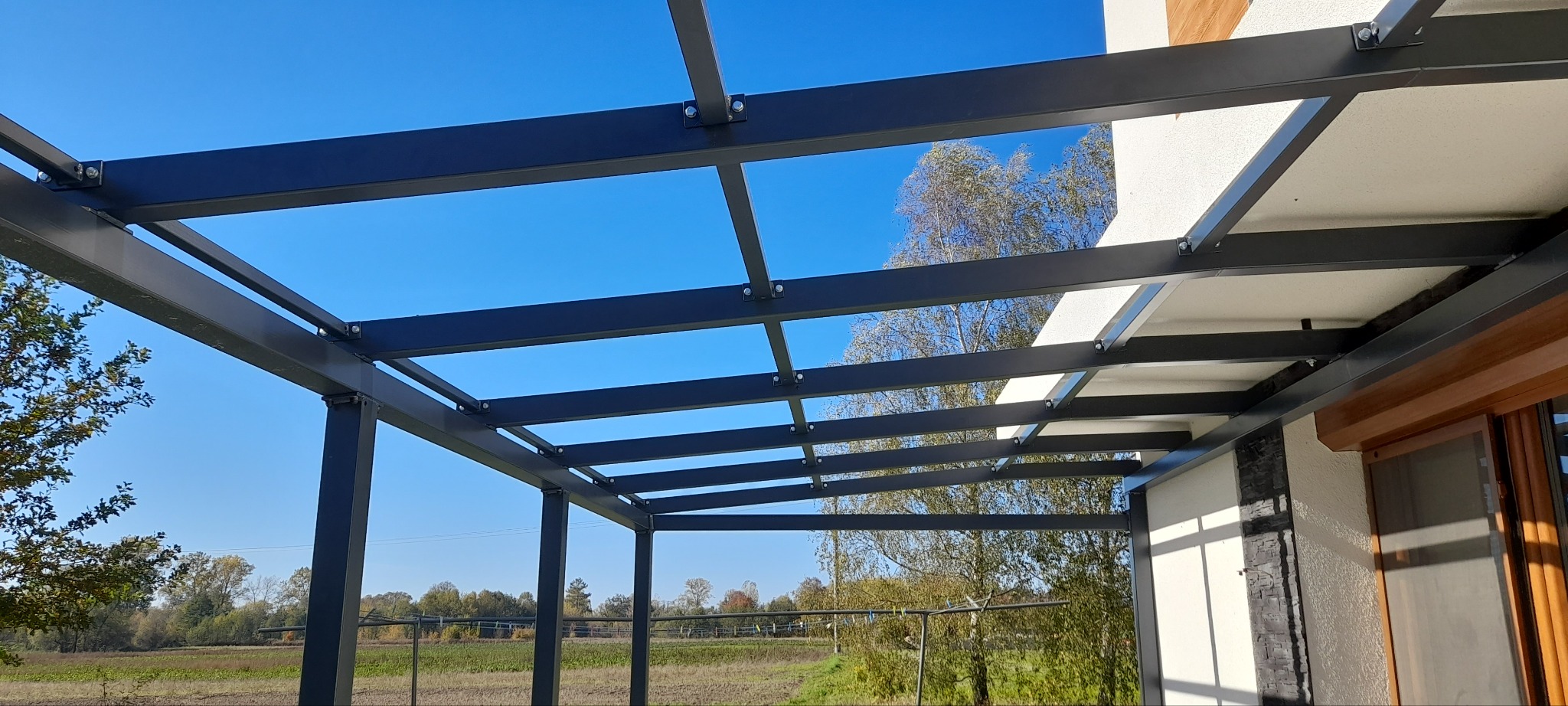 Nowoczesna, grafitowa pergola z metalową konstrukcją, widok z dołu na tle błękitnego nieba i zieleni. Konstrukcja przymocowana do budynku.
