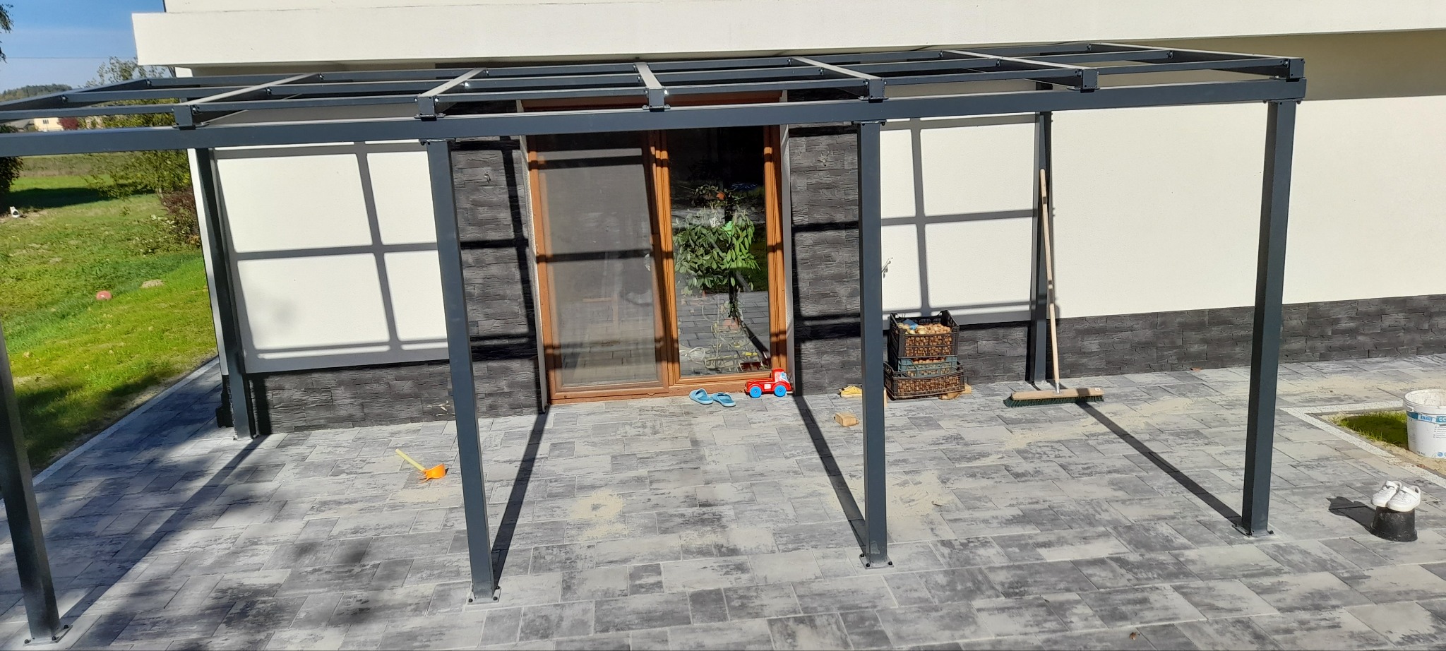 Metalowa konstrukcja zadaszenia tarasu w trakcie montażu, widoczne słupy i elementy mocujące, kostka brukowa na podłożu.