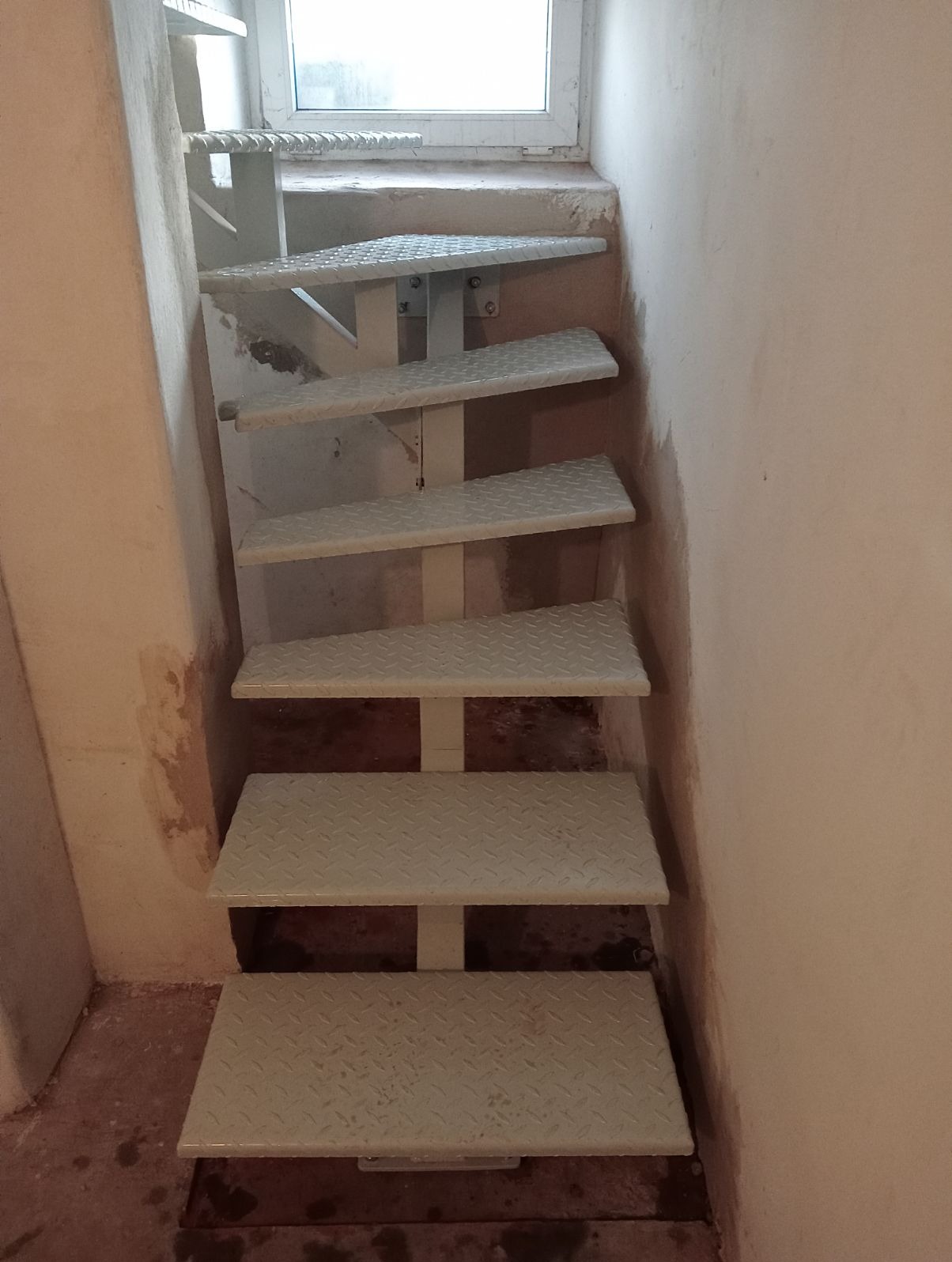 Metalowe schody spiralne o ażurowej konstrukcji, prowadzące w górę w wąskiej przestrzeni w pobliżu okna, surowy styl, widoczne ślady prac wykończeniowych na ścianach.