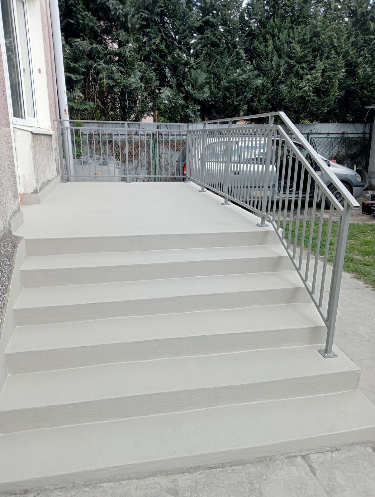 Nowoczesne schody zewnętrzne z szarą, metalową balustradą, prowadzące na mały taras. W tle zieleń drzew i zaparkowany samochód.