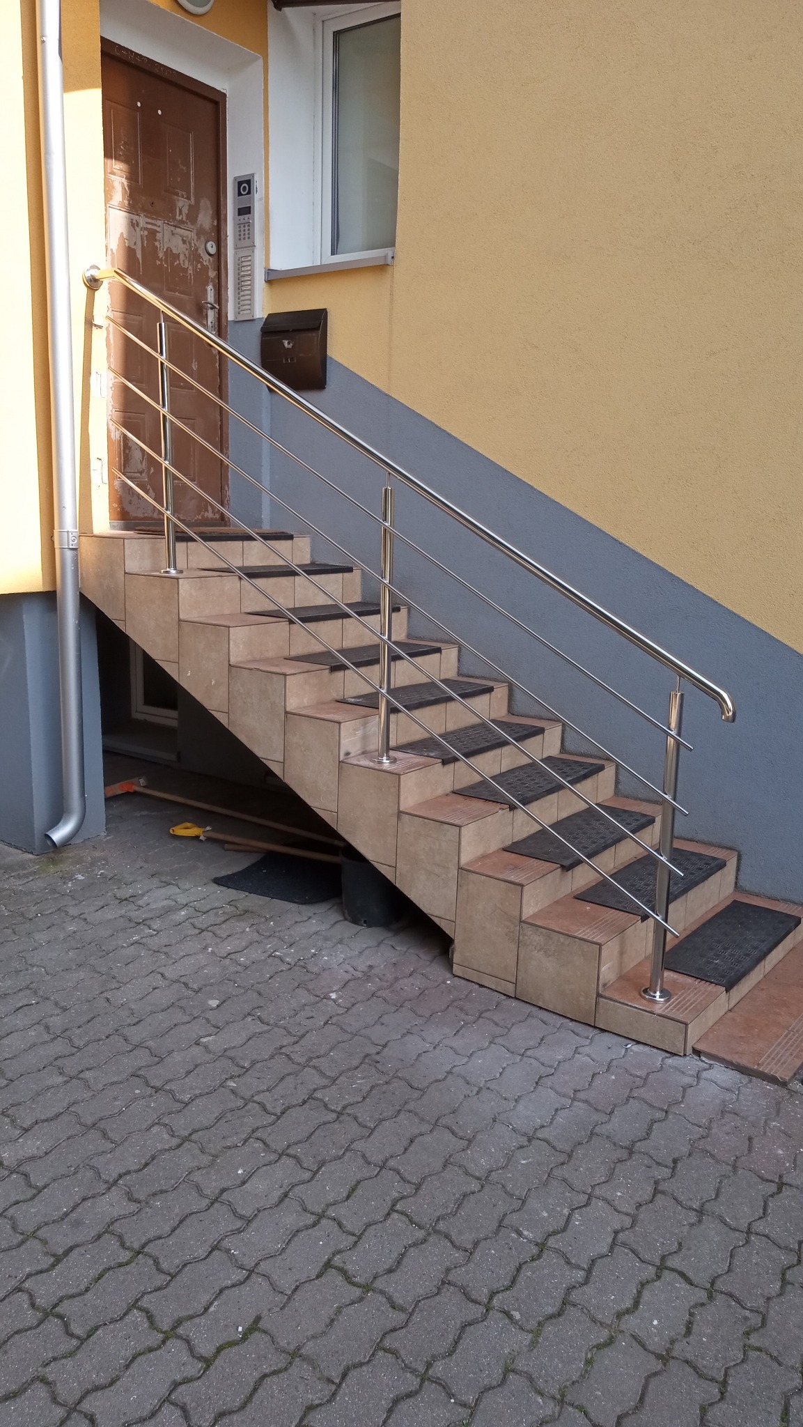 Zewnętrzne schody z beżowych płytek z czarnymi wycieraczkami, zabezpieczone błyszczącą, nierdzewną balustradą, prowadzące do drzwi wejściowych.