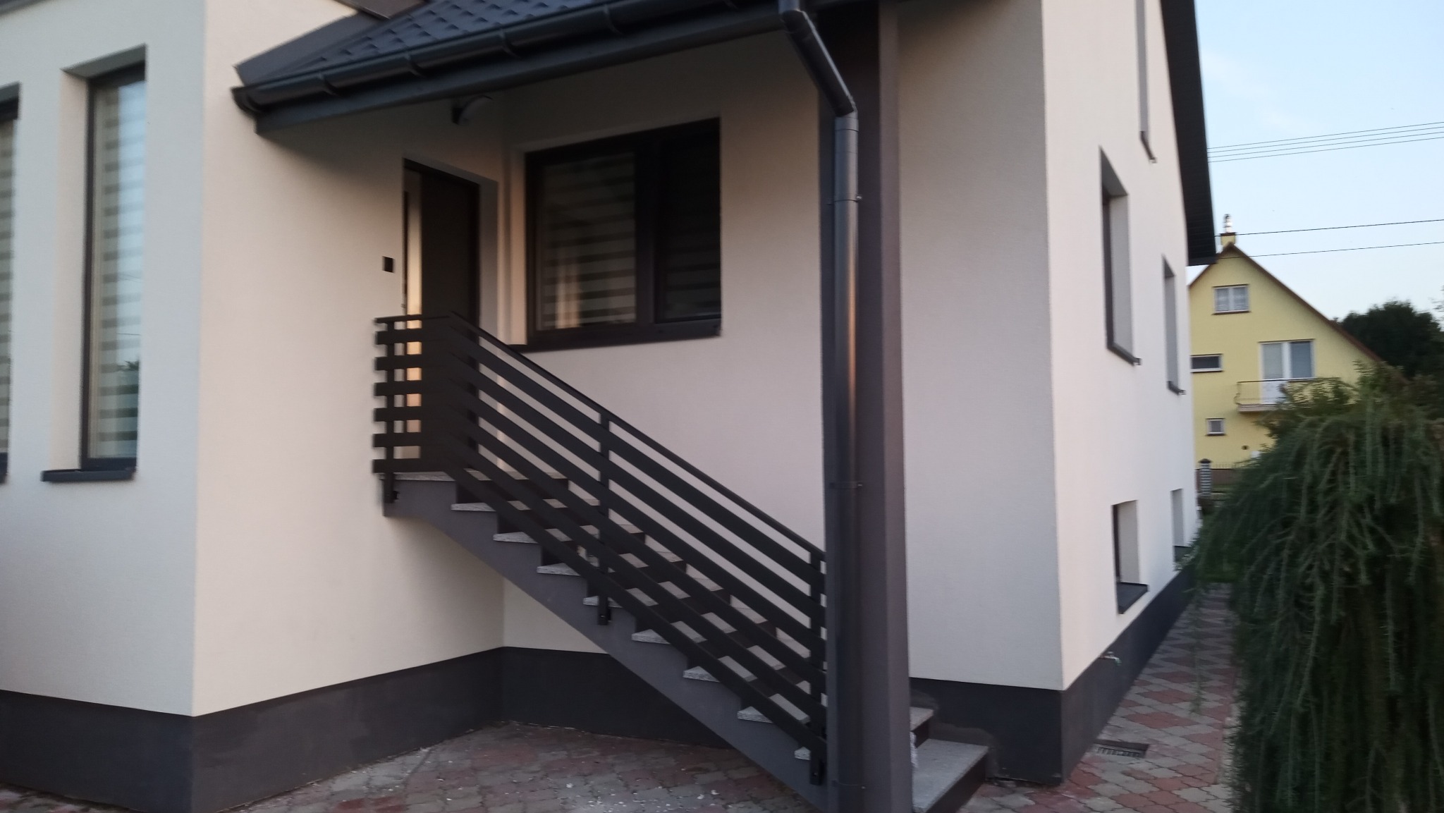 Zewnętrzne schody z ciemną, metalową balustradą prowadzące do drzwi wejściowych w jasnym budynku. Widoczny fragment elewacji i okna z żaluzjami.