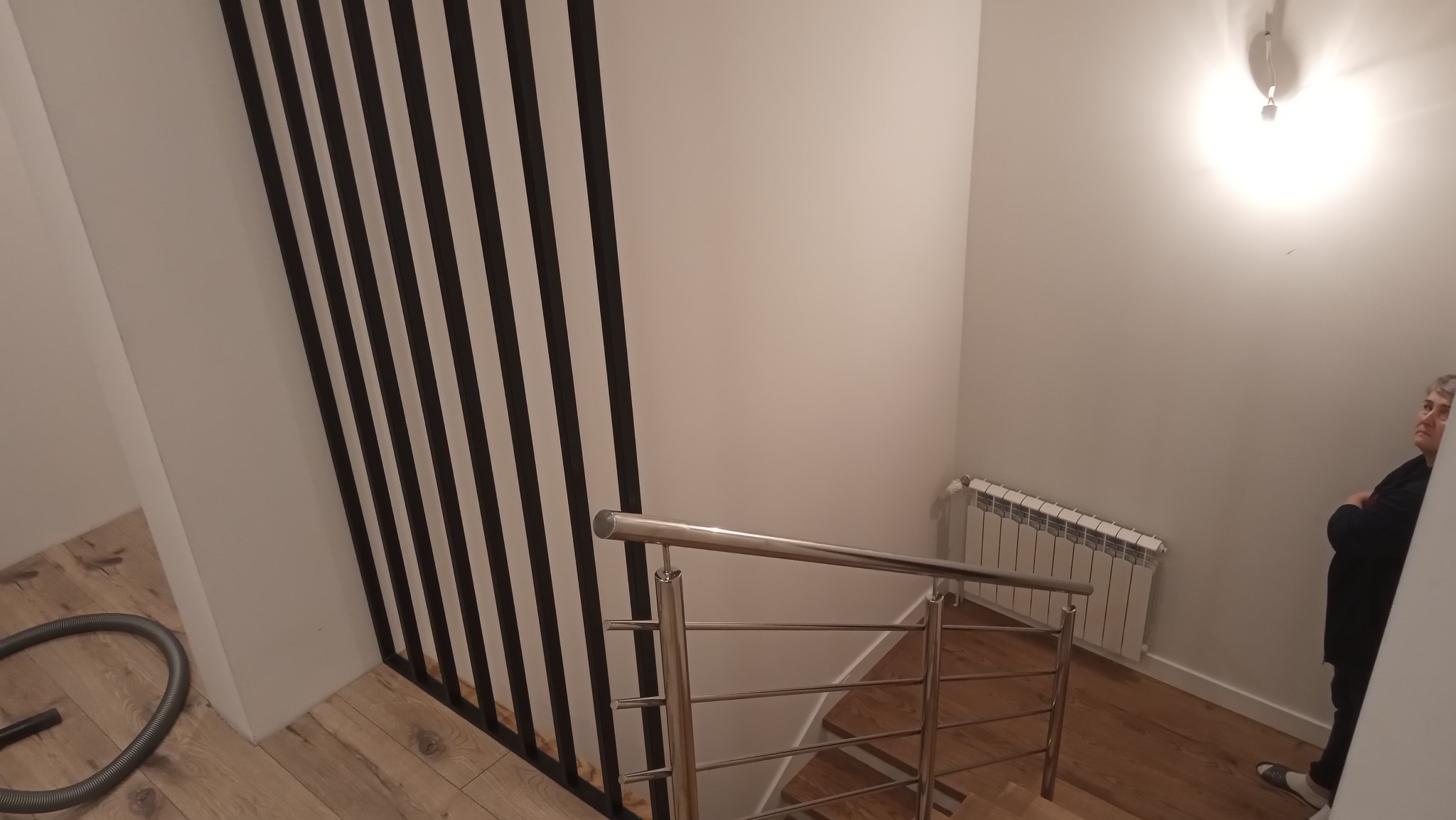 Nowoczesne schody z metalową balustradą i czarnymi, pionowymi elementami dekoracyjnymi. Widoczny fragment odkurzacza i osoba w tle.
