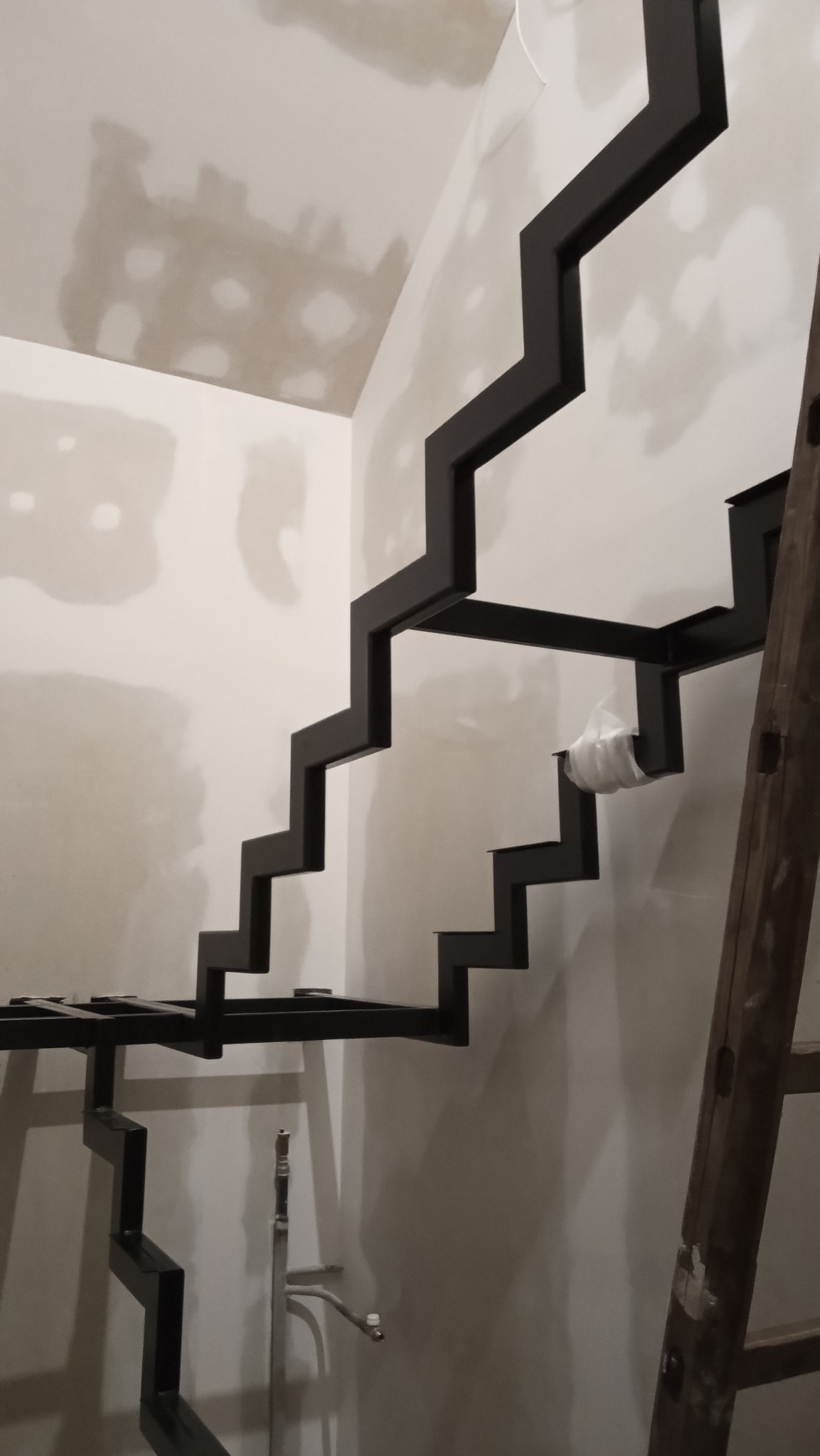 Nowoczesna, ażurowa konstrukcja schodów metalowych w surowym wnętrzu. Czarna, geometryczna forma na tle białych ścian, drabina oparta o ścianę.