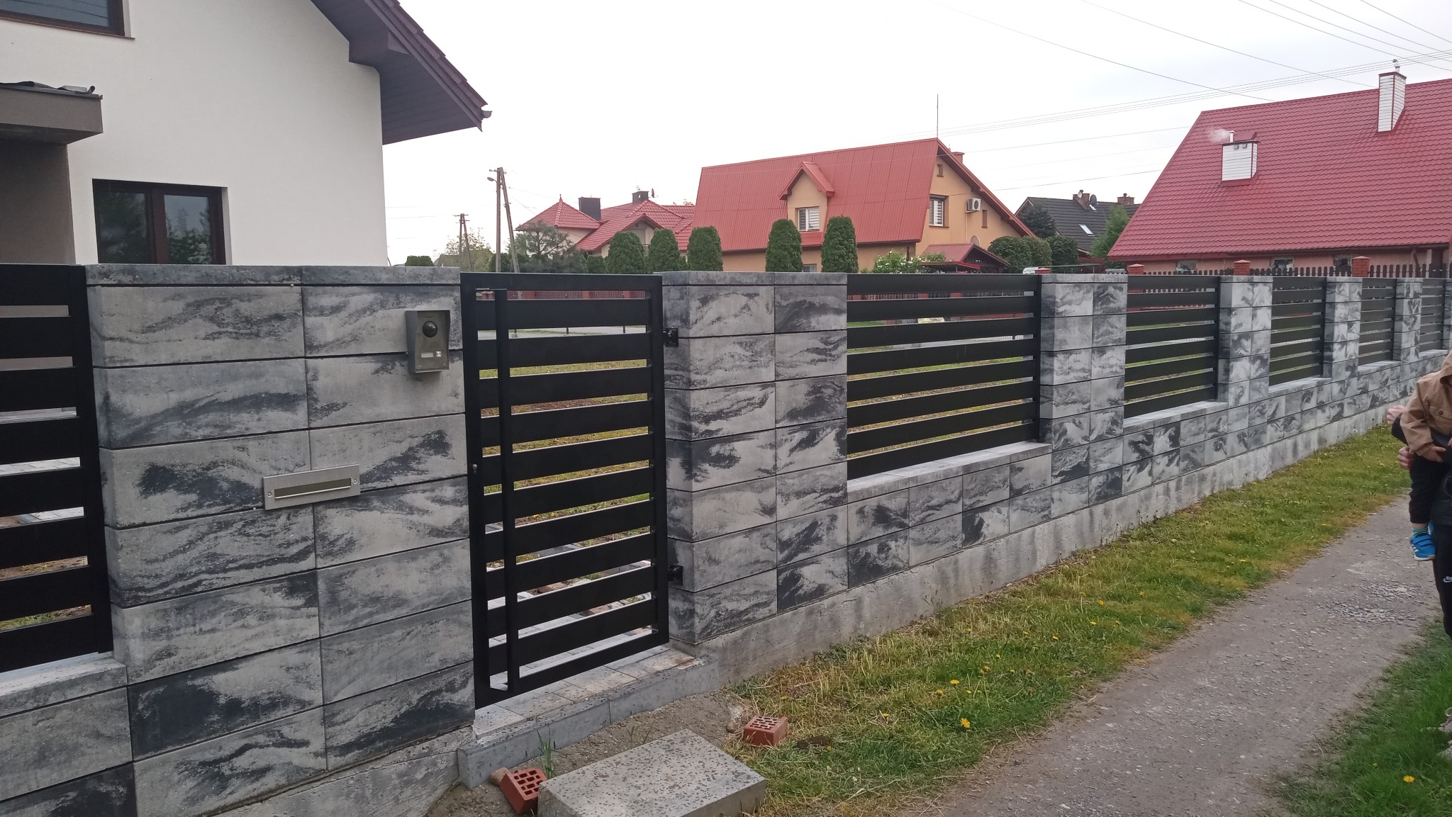 Nowoczesne ogrodzenie z szarej kostki i czarnych, poziomych przęseł, z furtką i skrzynką na listy, na tle domów z czerwonymi dachami.