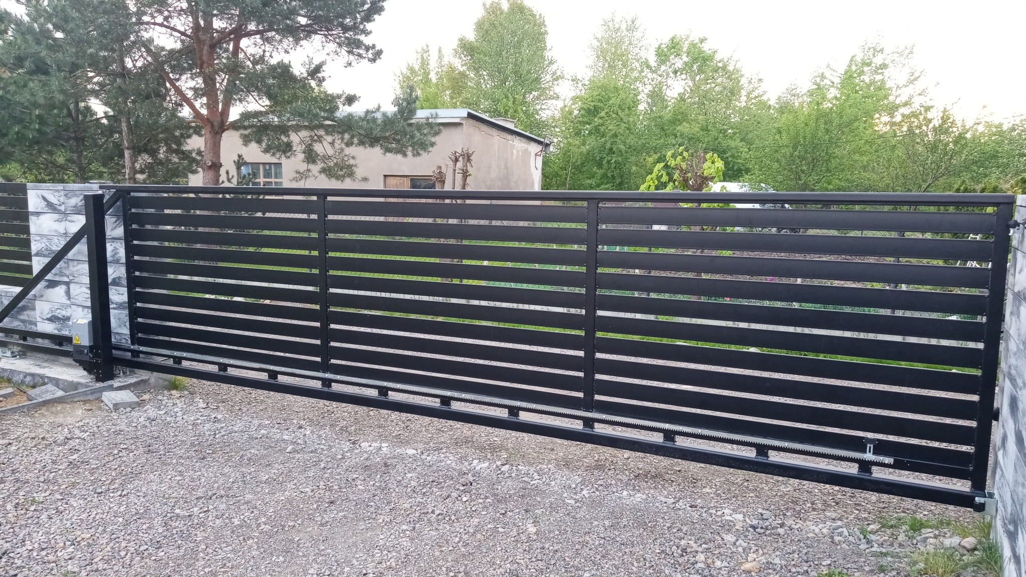 Automatyczna brama przesuwna z poziomymi profilami, czarna, na tle zieleni i budynku. Widoczny napęd i słupki z szarą okładziną. Żwirowe podłoże.
