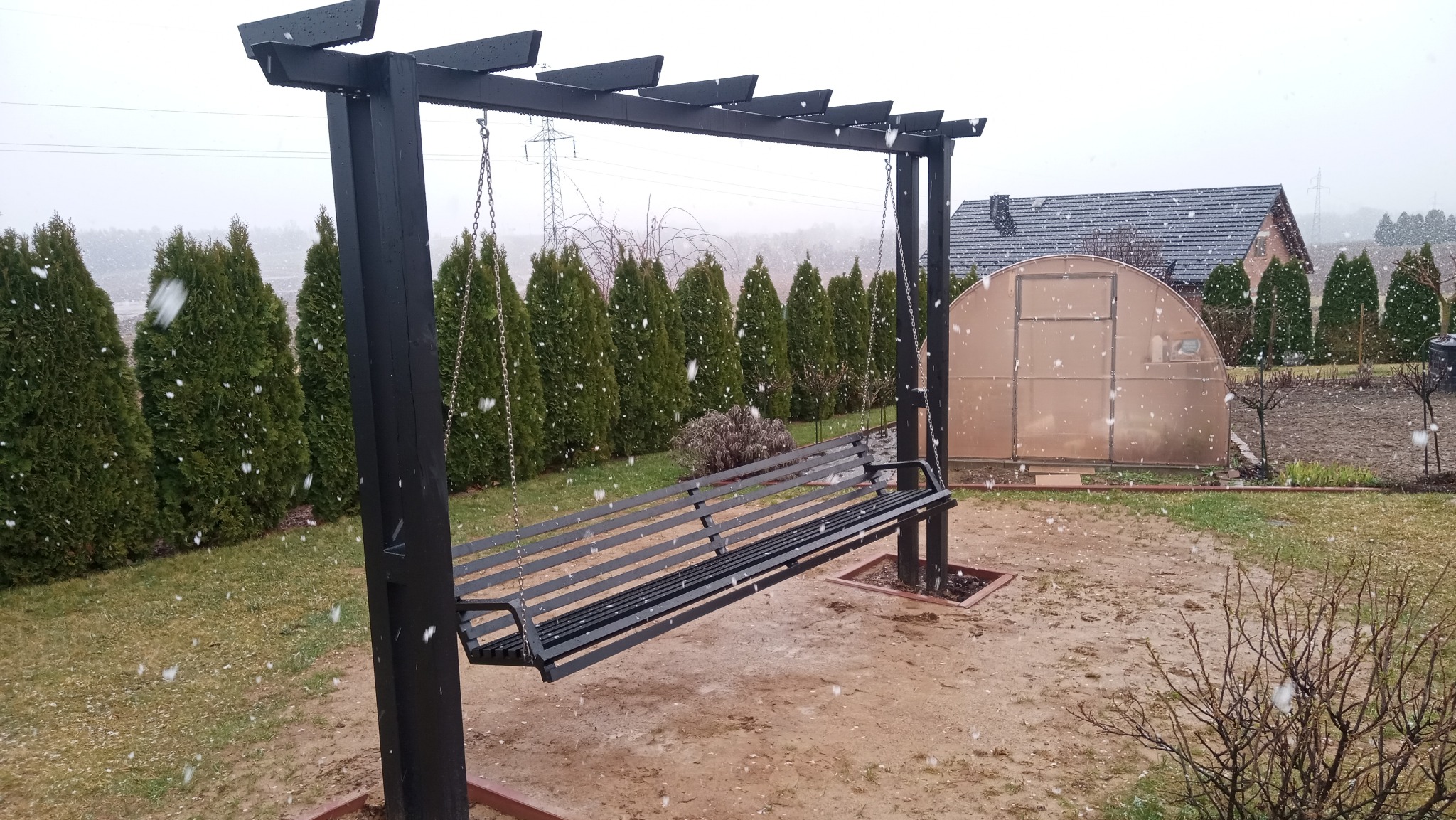 Czarna, metalowa huśtawka ogrodowa z łańcuchami, w tle rząd zielonych tuj i szklarnia, podczas opadów śniegu.