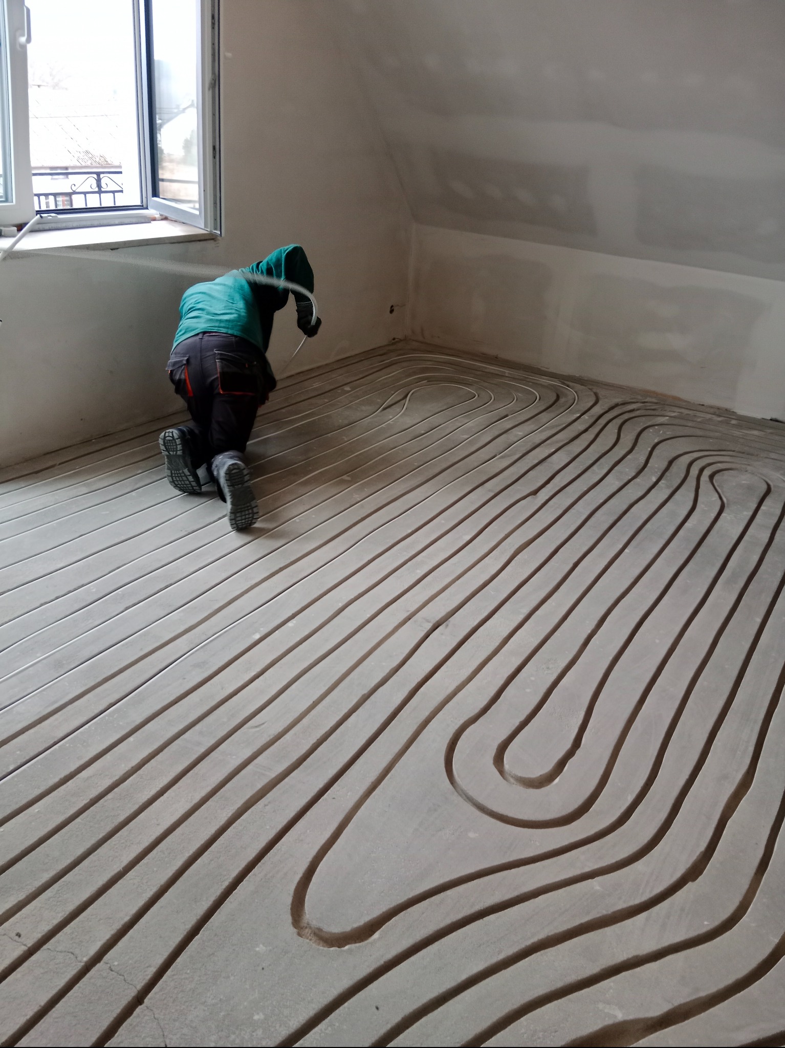 Instalator na kolanach montuje rury ogrzewania podłogowego w surowym wnętrzu z oknem. Widoczne pętle rur ułożone na podłożu, przygotowane do zalania wylewką.