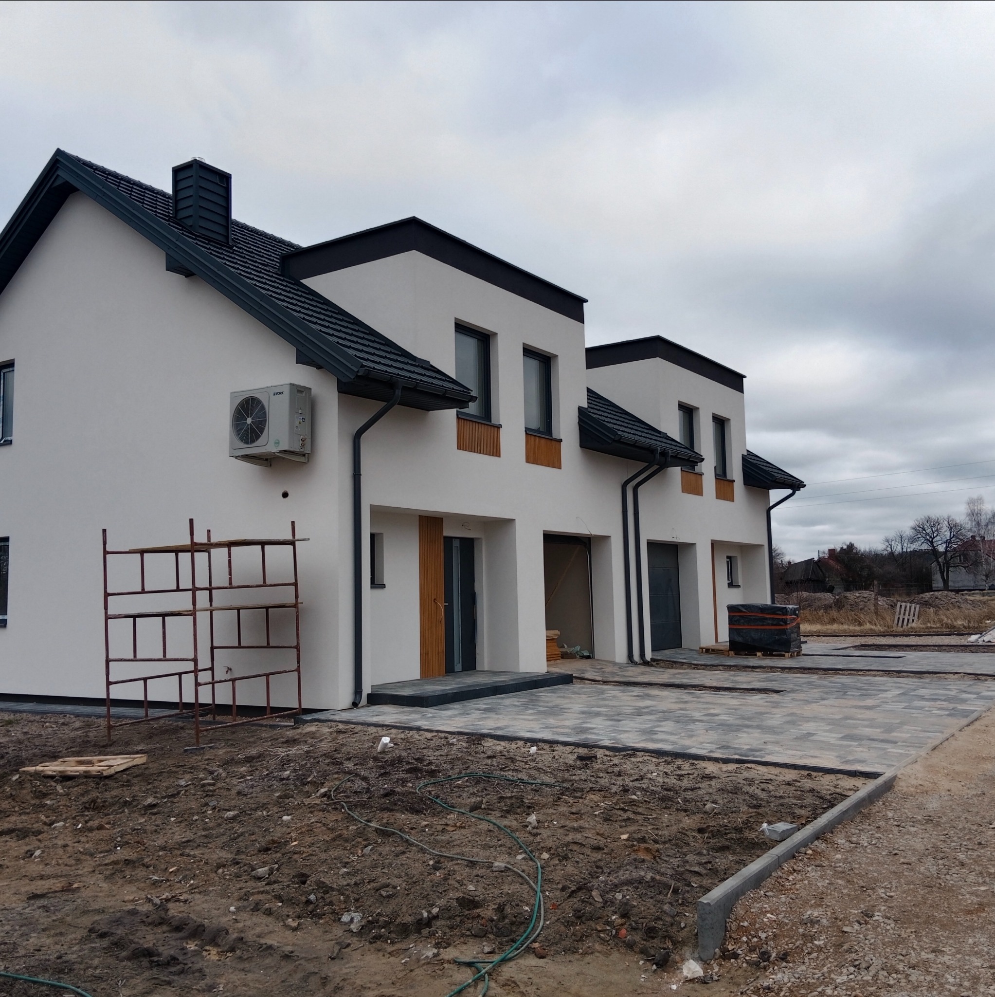 Nowoczesne, białe bliźniaki z klimatyzacją na elewacji, czarną dachówką i kostką brukową przed wejściem. Widoczne rusztowania i materiały budowlane.