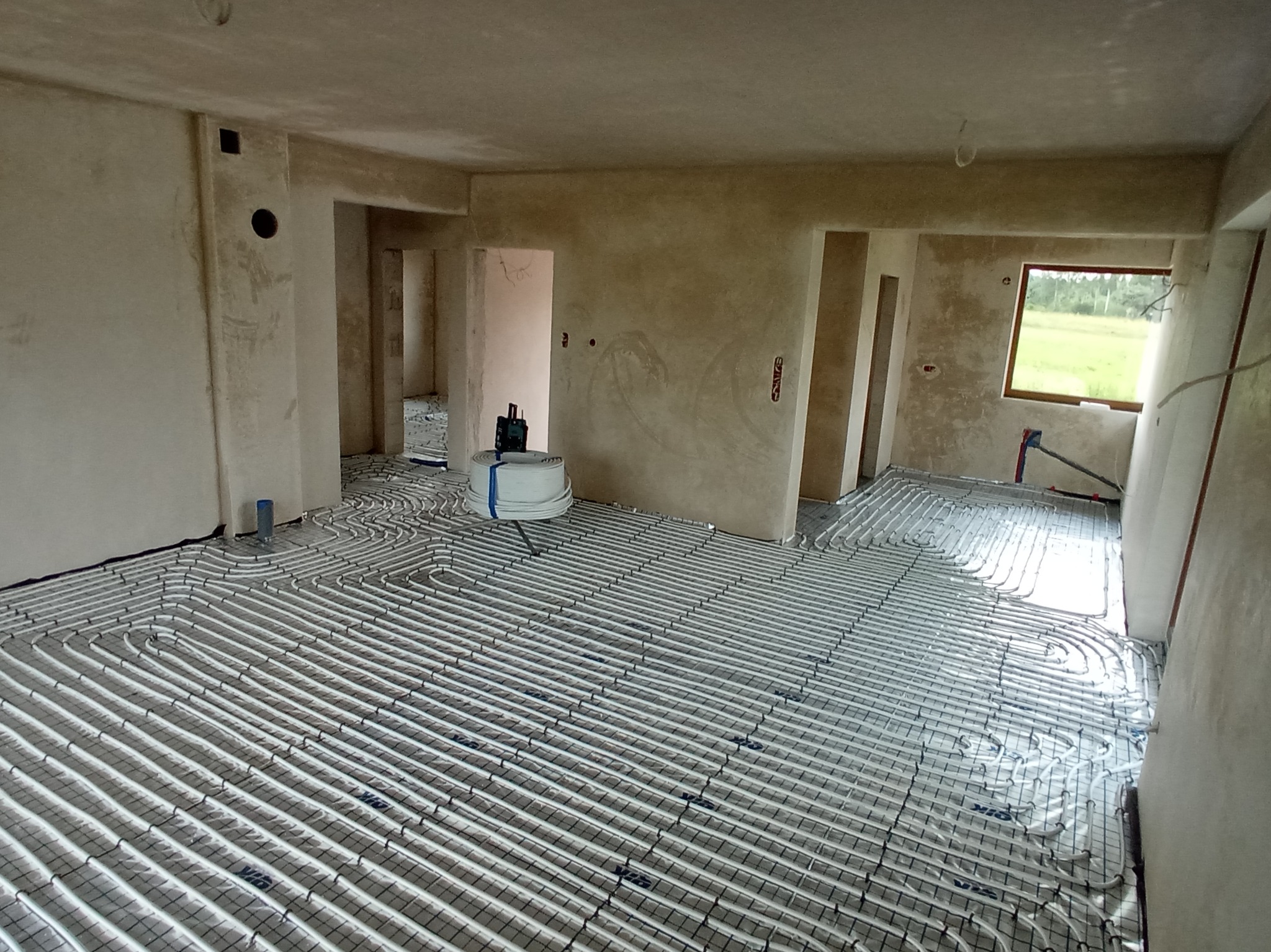 Rozłożona instalacja ogrzewania podłogowego w surowym wnętrzu, widoczne rury na siatce montażowej, przygotowane do zalania posadzką. W tle okno z widokiem na zieleń.