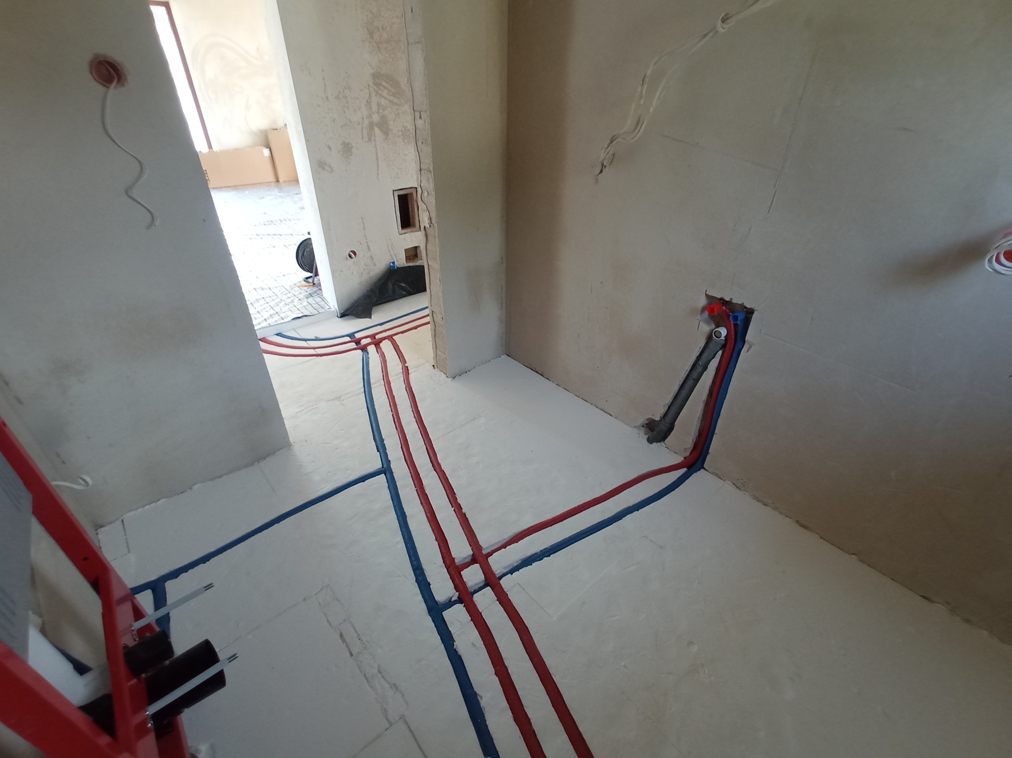 Instalacja wodna w nowym budynku: czerwone i niebieskie rury prowadzone po podłodze, wychodzące ze ściany, z widocznym stelażem podtynkowym WC. Surowy stan wykończenia.