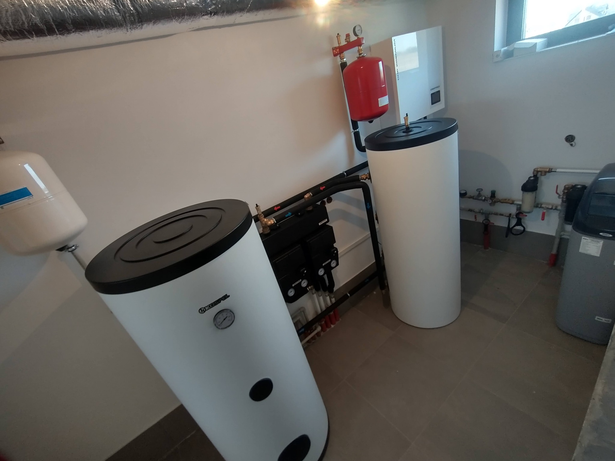Białe zbiorniki akumulacyjne ciepła w kotłowni, z widoczną instalacją hydrauliczną i rurami. Elementy kotłowni w pomieszczeniu z oknem i wentylacją.