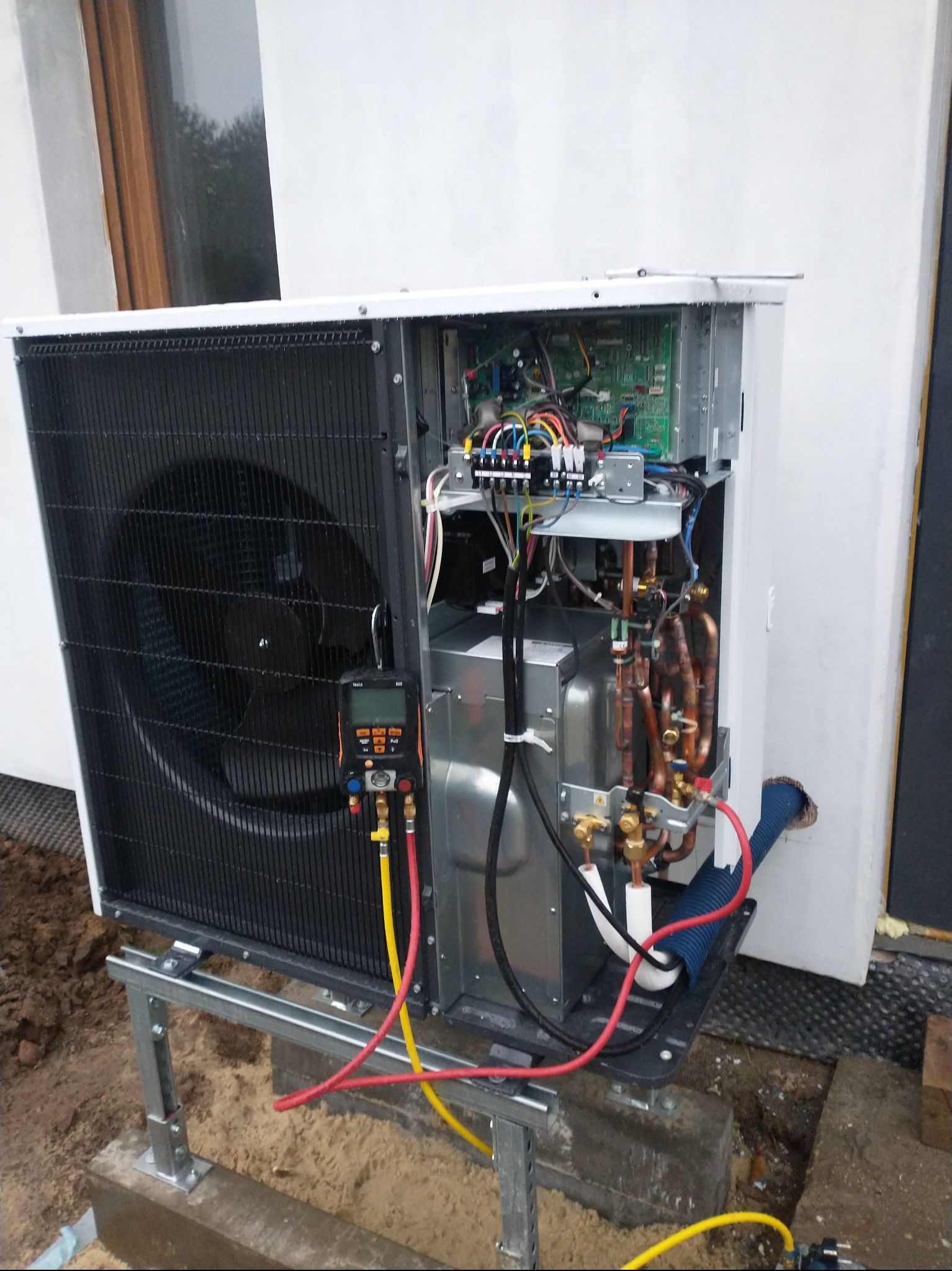 Otwarta pompa ciepła na zewnątrz budynku, widoczne elementy instalacji, wentylator i podłączenia. Pomiar ciśnienia za pomocą manometru.