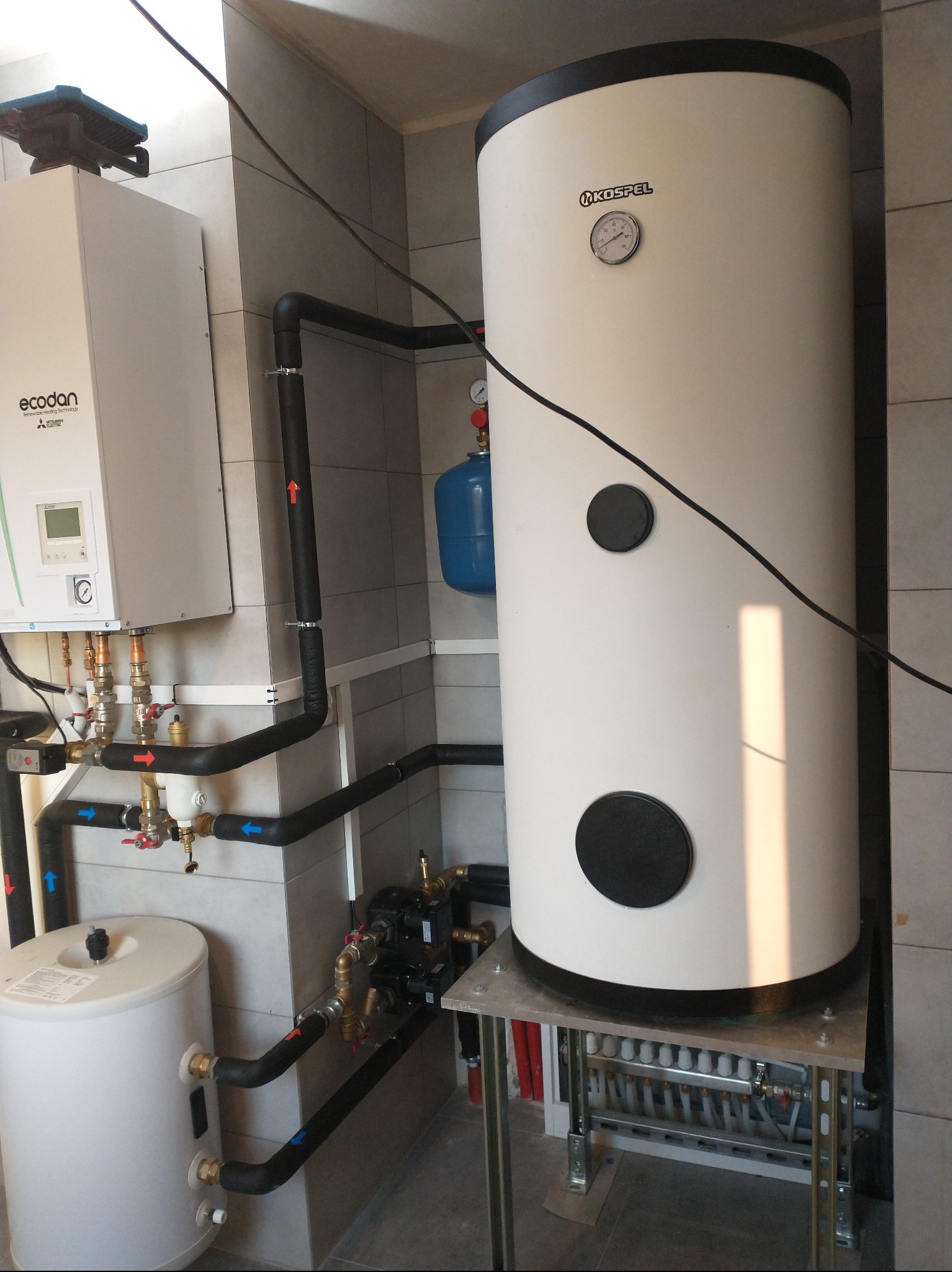 Biały zasobnik ciepłej wody Kospel z czarnymi zaślepkami, obok pompa ciepła Ecodan Mitsubishi i niebieski zbiornik wyrównawczy, połączone rurami w kotłowni.