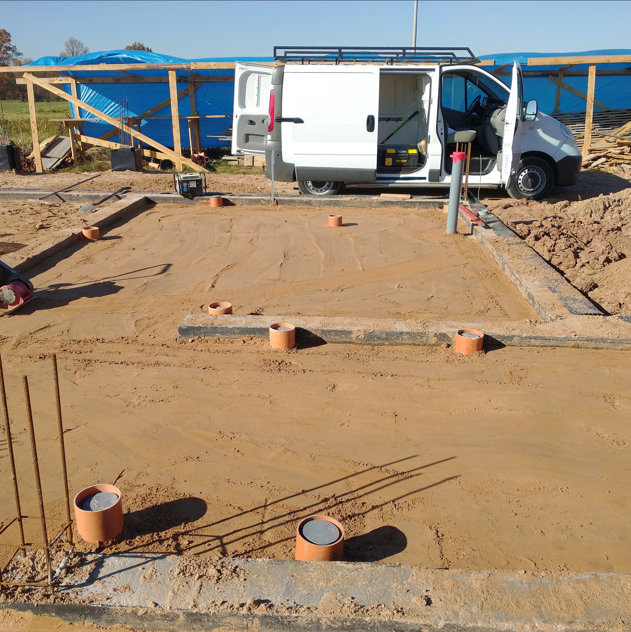 Przygotowanie terenu pod budowę: fundamenty z rurami kanalizacyjnymi, widoczny samochód dostawczy z otwartymi drzwiami, teren budowy w słoneczny dzień.