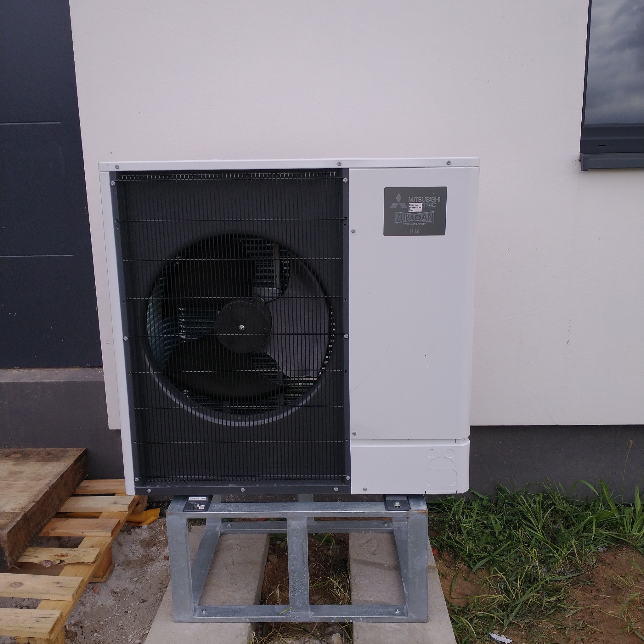 Biała pompa ciepła Mitsubishi Zubadan R32, zamontowana na metalowej konstrukcji przy ścianie budynku. Widoczne elementy instalacji i otoczenie.