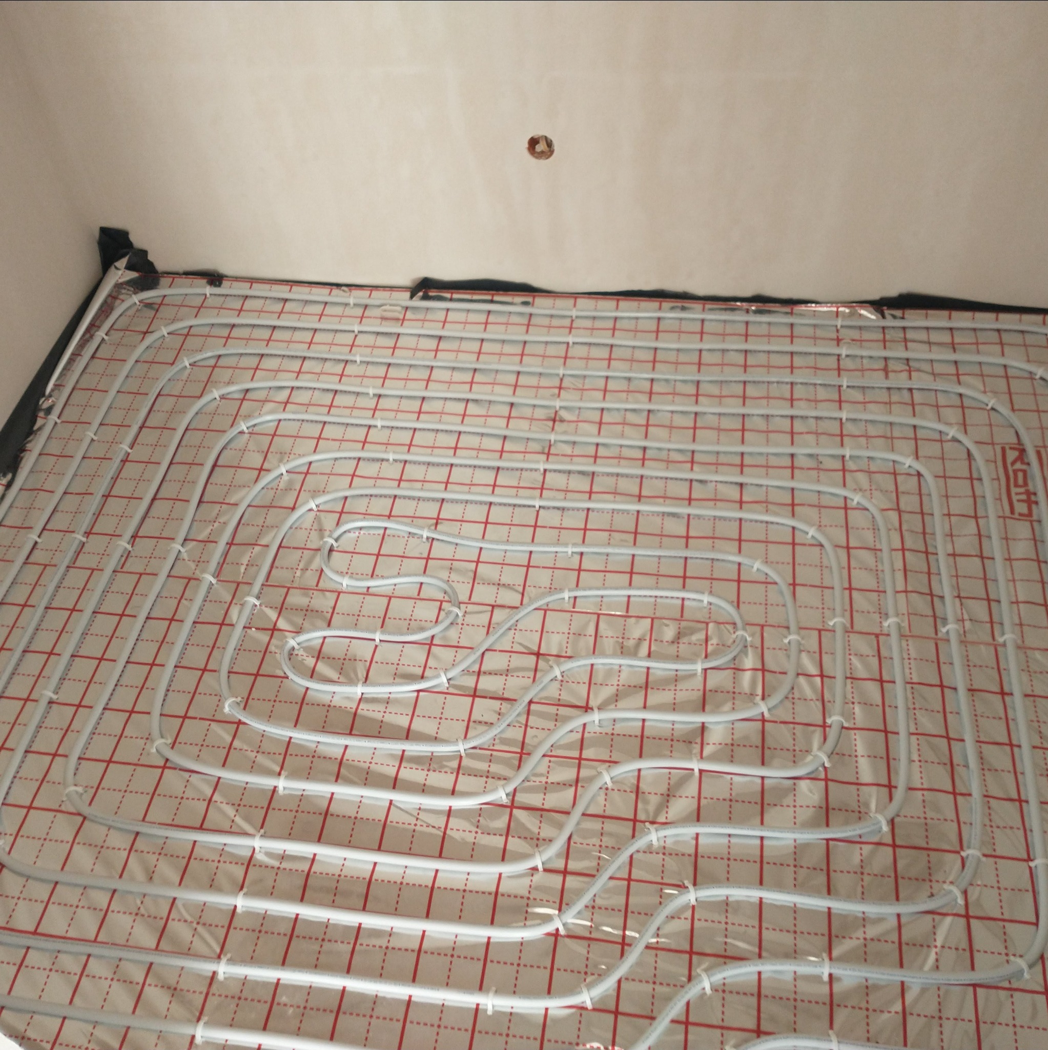 Ułożone rury ogrzewania podłogowego na folii z czerwoną siatką, zabezpieczone czarną taśmą przy ścianie, gotowe do zalania posadzką.