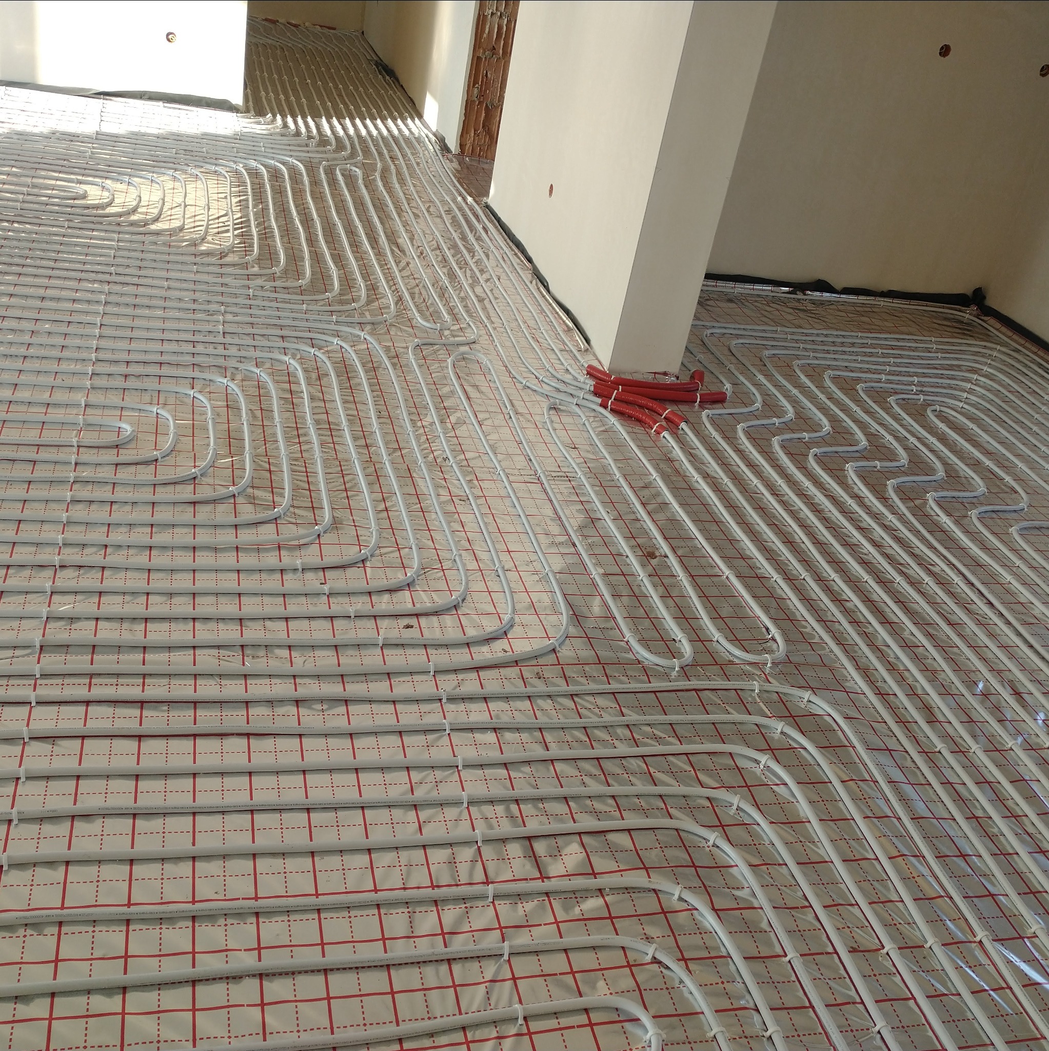 Białe rury ogrzewania podłogowego ułożone na folii z czerwoną siatką, widoczne fragmenty ścian i czerwone rury przyłączeniowe. Ujęcie z góry, perspektywa.