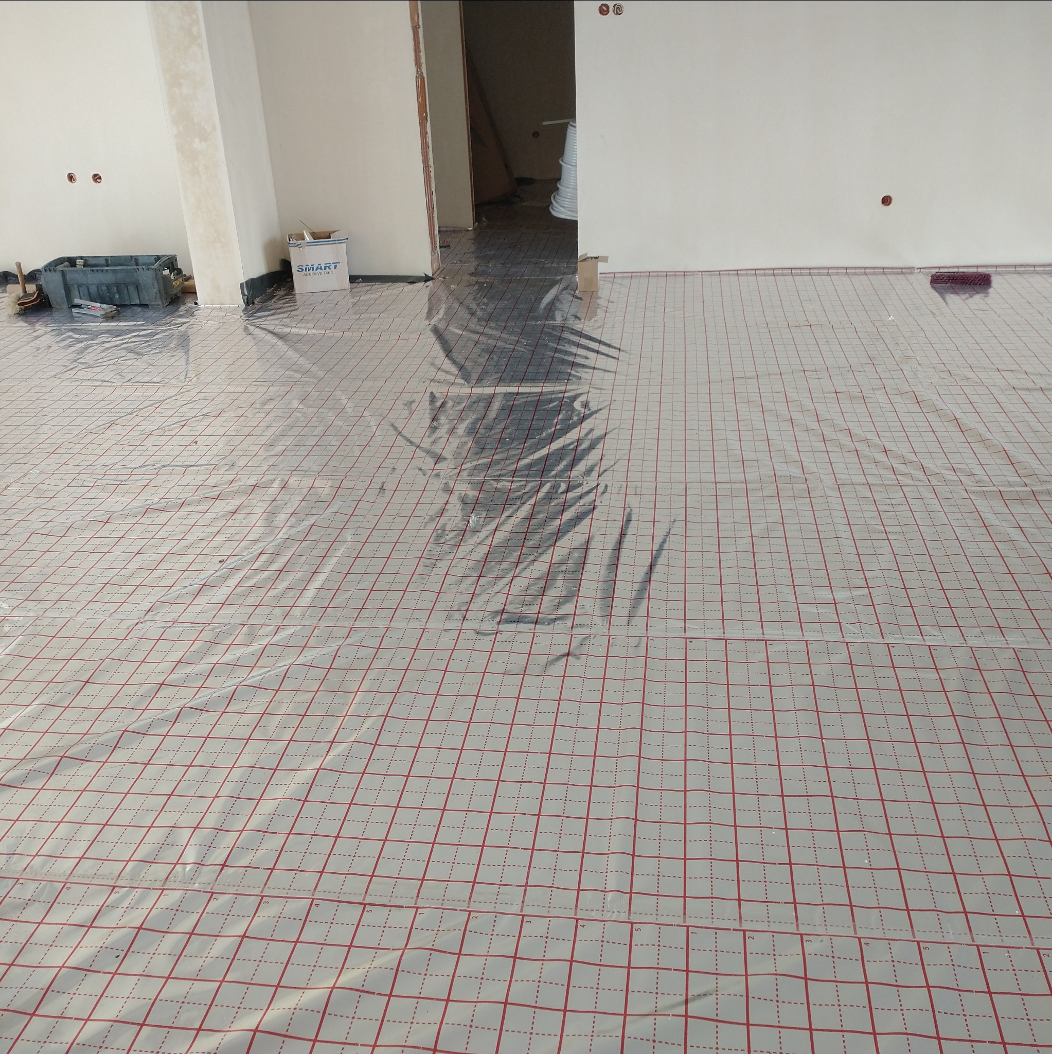 Rozłożona folia z siatką na podłodze, przygotowana pod ogrzewanie podłogowe w surowym wnętrzu. Widoczne narzędzia i materiały budowlane w tle.