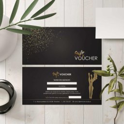 CLICK MADE KAROLINA WCISŁO - Elegancki czarno-złoty voucher prezentowy z danymi kontaktowymi na drewnianym tle, ozdobiony roślinami i świecą, sugerujący luksusową ofertę.