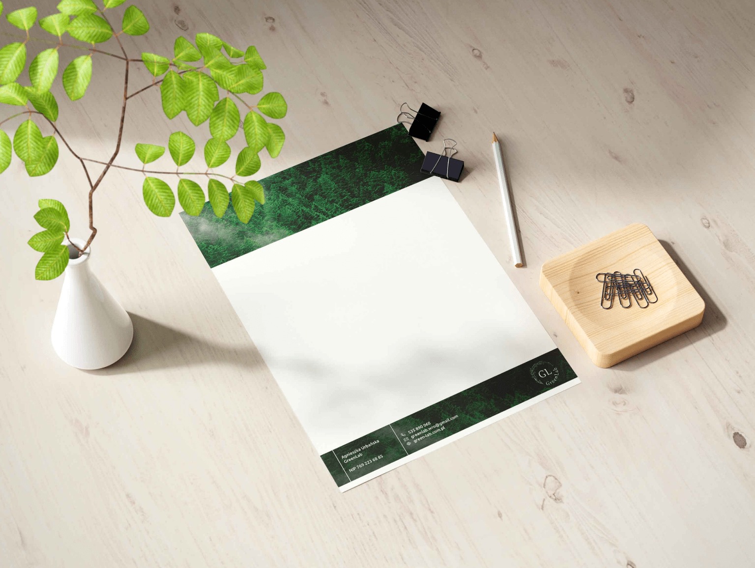Papier firmowy GreenLab z zielonym motywem leśnym, leżący na biurku obok białego wazonu z gałązką, drewnianego pojemnika na spinacze i ołówka. Minimalistyczny design.