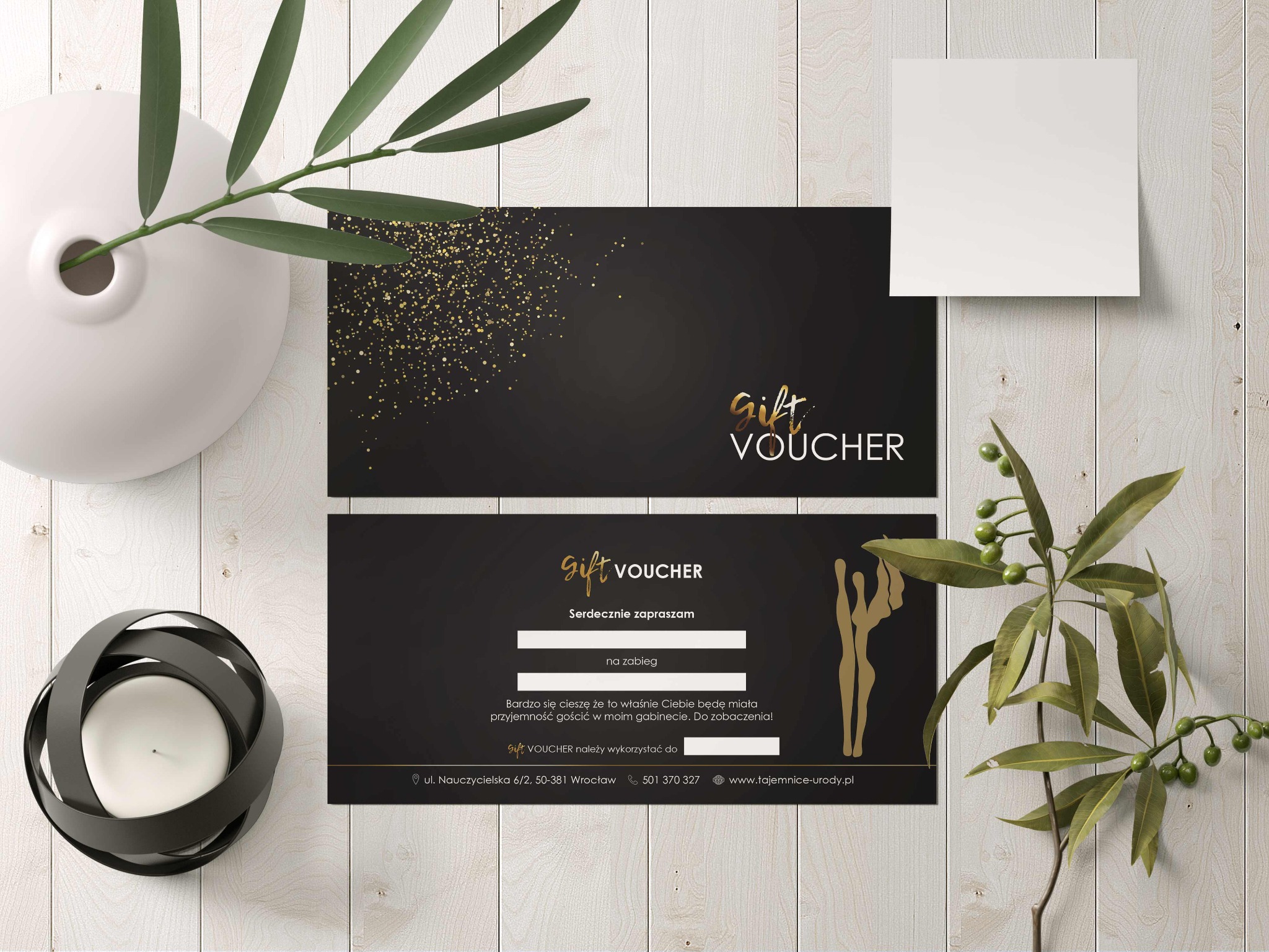 Elegancki czarno-złoty voucher prezentowy z danymi kontaktowymi na drewnianym tle, ozdobiony roślinami i świecą, sugerujący luksusową ofertę.