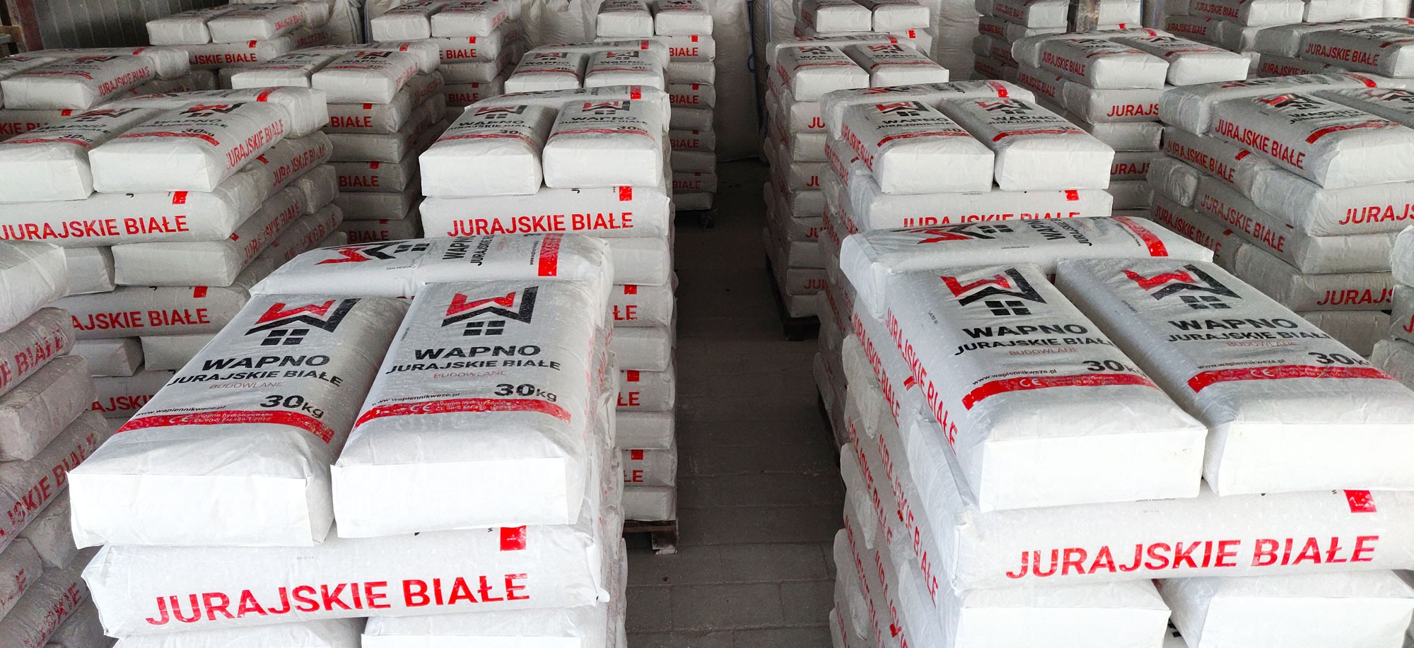 Magazyn z paletami białego wapna budowlanego 'Jurajskie Białe' w workach 30kg. Widok na rzędy ułożonych palet z bliska, z widocznym logo producenta na każdym worku.