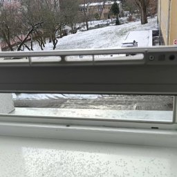 Przemysław Domański - Białe okno PCV z widokiem na zimowy krajobraz. Widoczny mechanizm rolety. Okno uchylone, na parapecie osad z białego pyłu. Ujęcie z bliska, wnętrze.