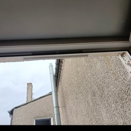 Przemysław Domański - Widok z okna PCV na elewację budynku z rynną spustową. Fragment ramy okiennej i uszczelki widoczne w kadrze.