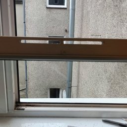 Przemysław Domański - Otwarty profil okna PCV z widocznym uszczelnieniem i elementami mocującymi. Widok na szary budynek z rynną i oknami w tle. Na parapecie narzędzia.