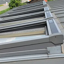 Szare rolety zewnętrzne na dachu z dachówki ceramicznej, widok z dołu pod kątem, fragment chodnika z pieszym w tle.