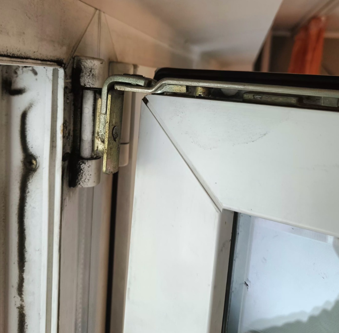 Detail uszkodzonego okna PCV: brudne, białe ramy, widoczne metalowe okucia i zawiasy. Widoczne ślady pleśni na oknie.