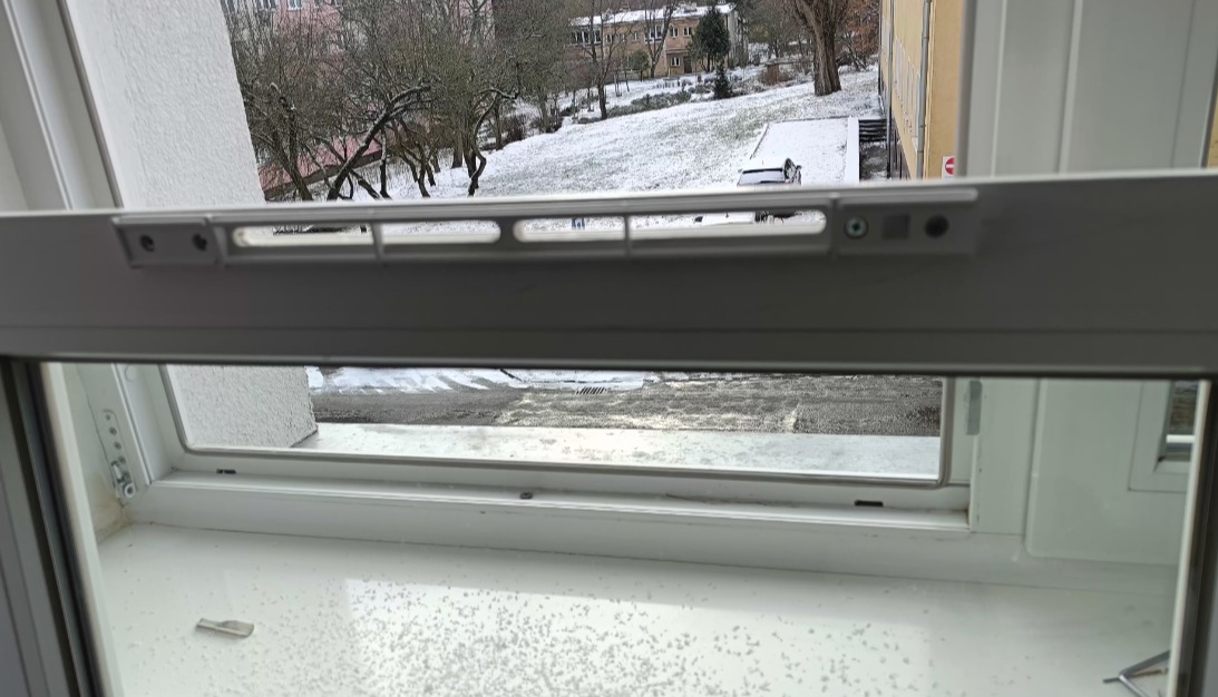 Białe okno PCV z widokiem na zimowy krajobraz. Widoczny mechanizm rolety. Okno uchylone, na parapecie osad z białego pyłu. Ujęcie z bliska, wnętrze.