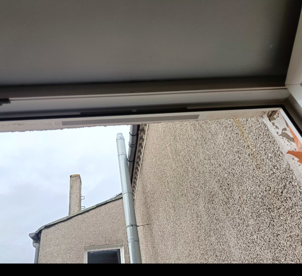 Widok z okna PCV na elewację budynku z rynną spustową. Fragment ramy okiennej i uszczelki widoczne w kadrze.