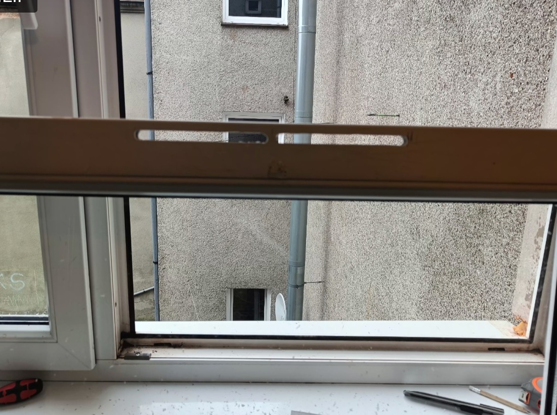 Otwarty profil okna PCV z widocznym uszczelnieniem i elementami mocującymi. Widok na szary budynek z rynną i oknami w tle. Na parapecie narzędzia.