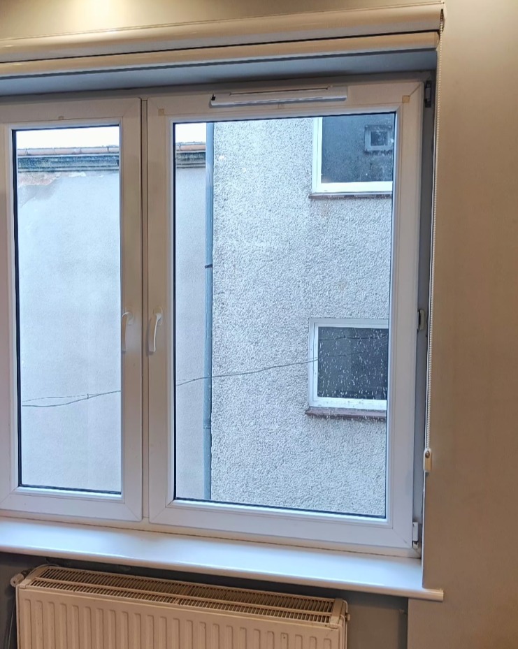 Białe okno PCV z roletą zwiniętą u góry, widok na szary budynek z oknami. Pod oknem grzejnik. Neutralne oświetlenie.