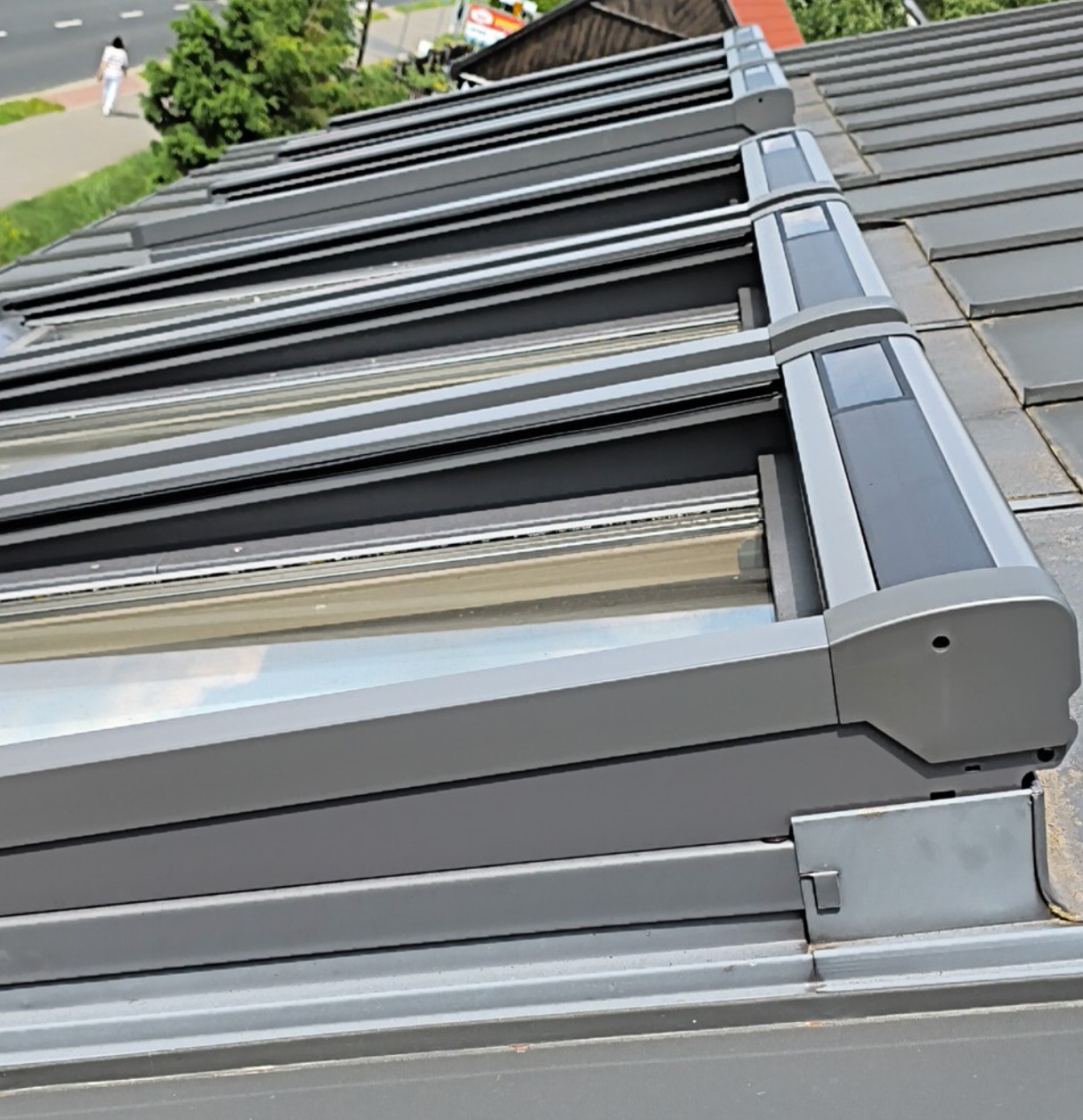 Szare rolety zewnętrzne na dachu z dachówki ceramicznej, widok z dołu pod kątem, fragment chodnika z pieszym w tle.