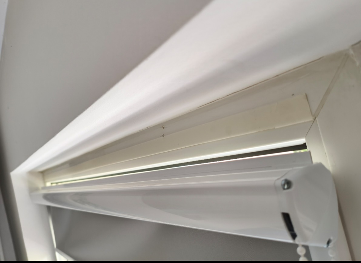 Zamknięta, biała roleta okienna w kasecie, zamontowana w oknie PCV. Widoczny mechanizm łańcuszkowy. Ujęcie z dołu, blisko rolety.