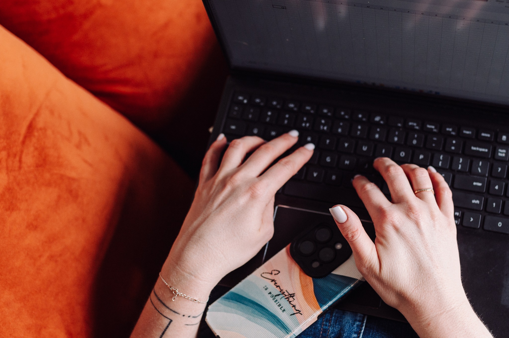 Kobieta pisze na laptopie; widoczny telefon z napisem 'Everything is possible', dłoń z bransoletką i tatuażem na tle pomarańczowej sofy.
